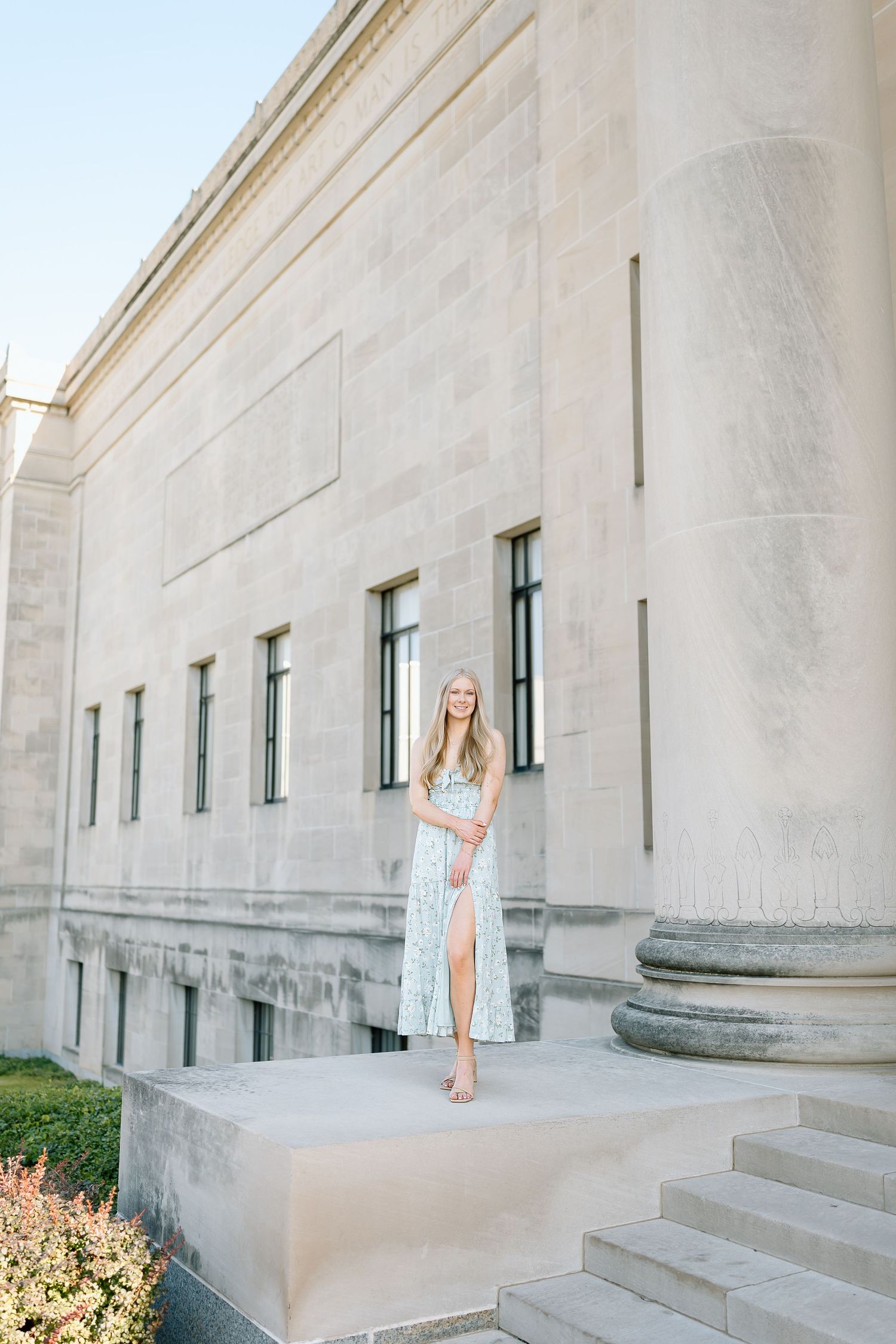 A person in a white dress poses near stone columns and steps of a grand building exterior.