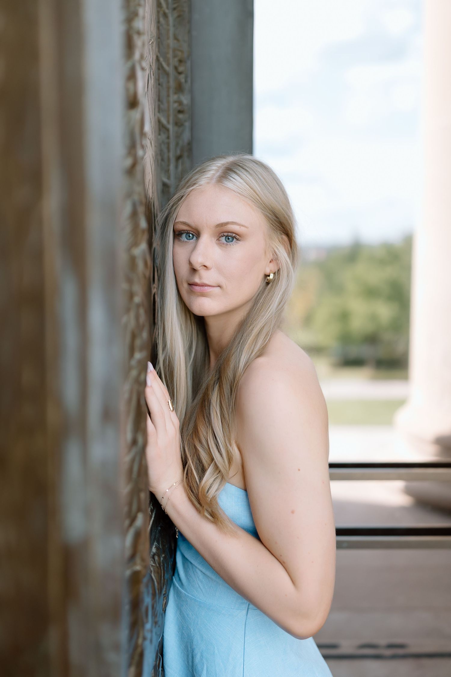 A person in a light blue top poses against wooden pillars in an outdoor setting.