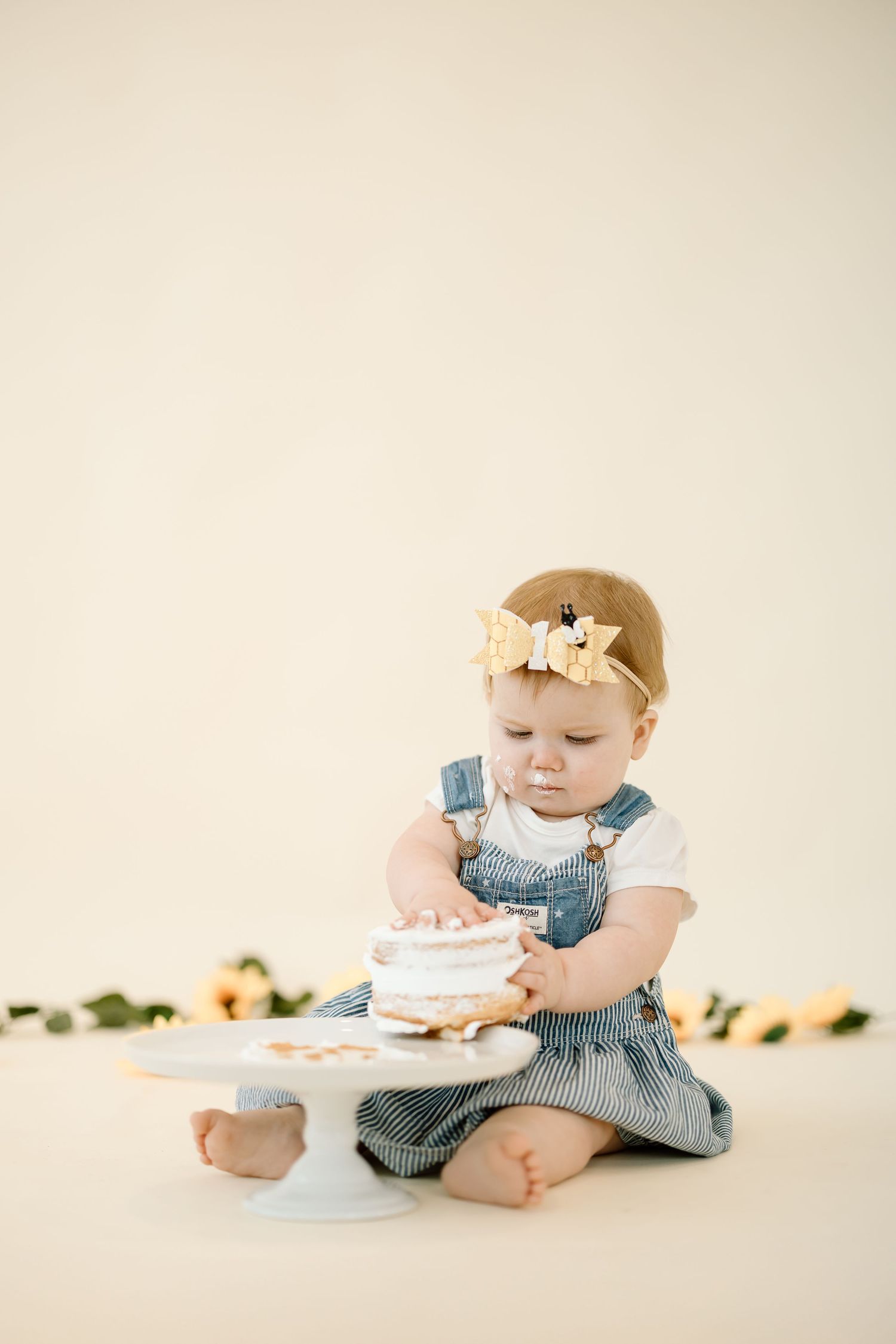 Baby in blue denim dress with yellow headband sits on cream background with cake stand.