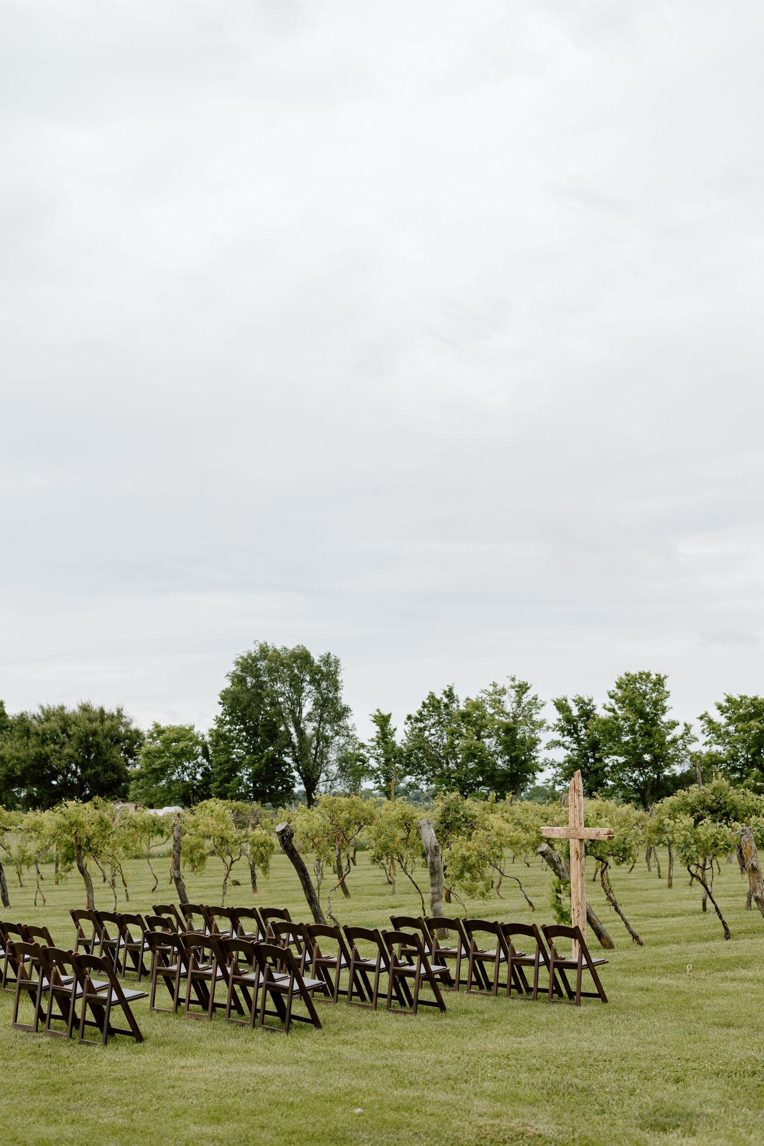Outdoor wedding ceremony setup with dark wooden folding chairs and a rustic wooden cross on a green lawn.