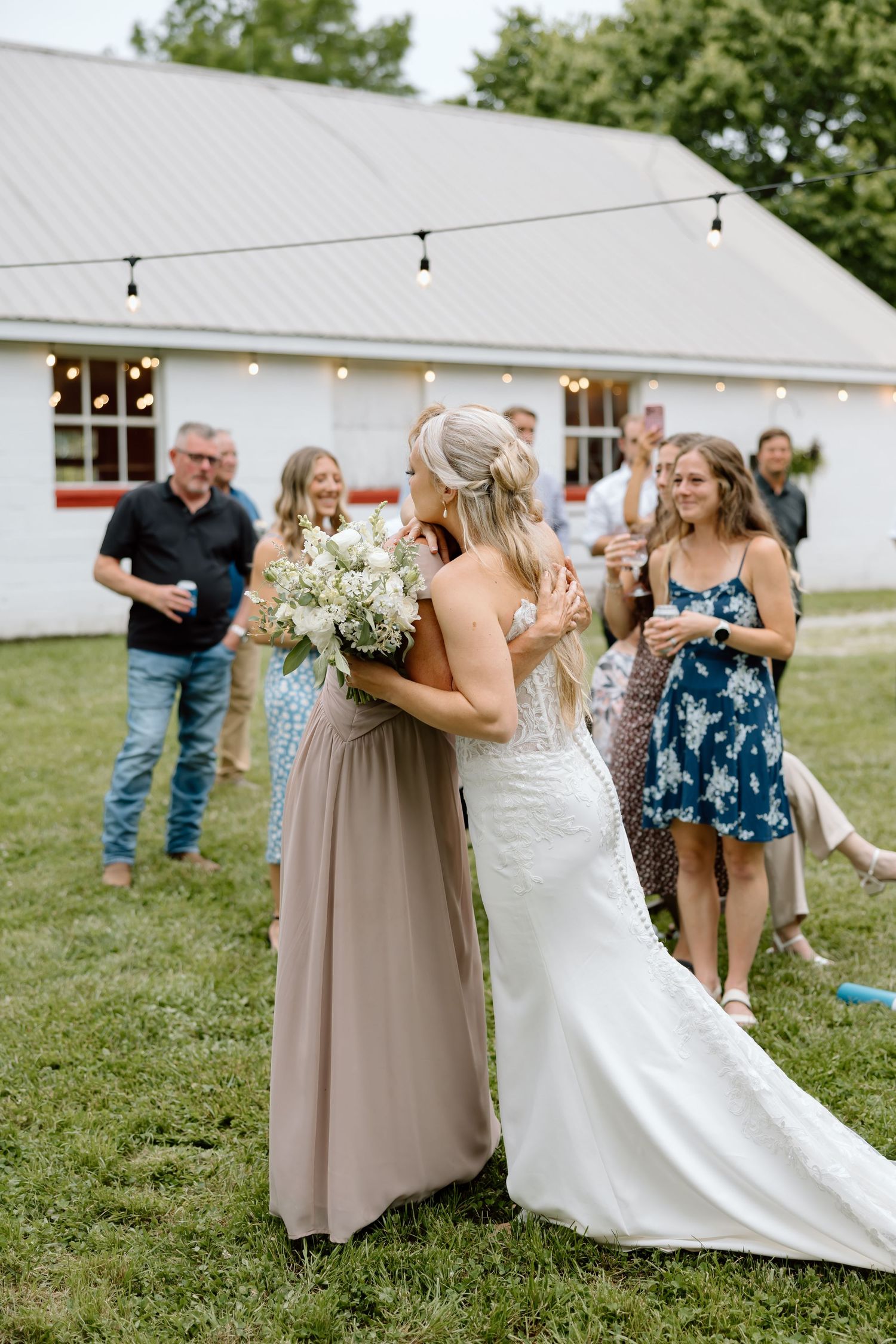 Bride in white lace gown hugging bridesmaid in taupe dress outdoors at rustic farm wedding venue with string lights.
