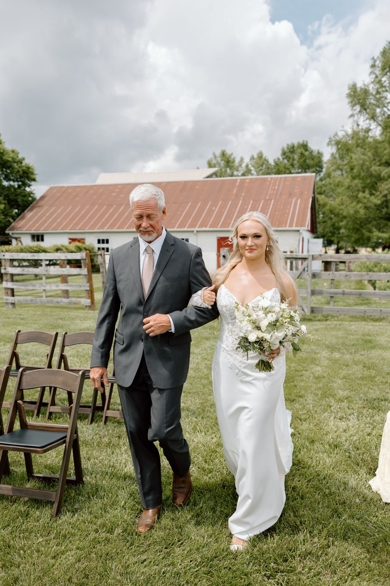 Bride in white gown walks down outdoor aisle with older man in gray suit at rustic farm wedding venue.