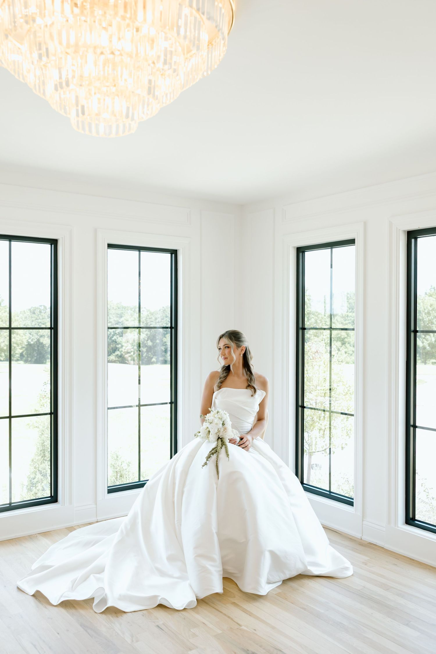 Bride in a strapless white ball gown holding a bouquet, standing in a bright white room with large windows and a chandelier.