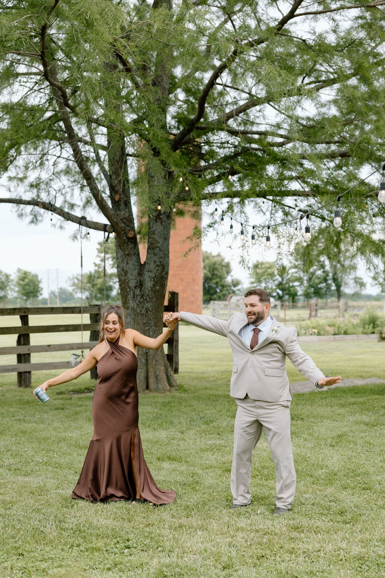 A couple dances outdoors on green grass near a large tree at a rustic farm wedding venue.