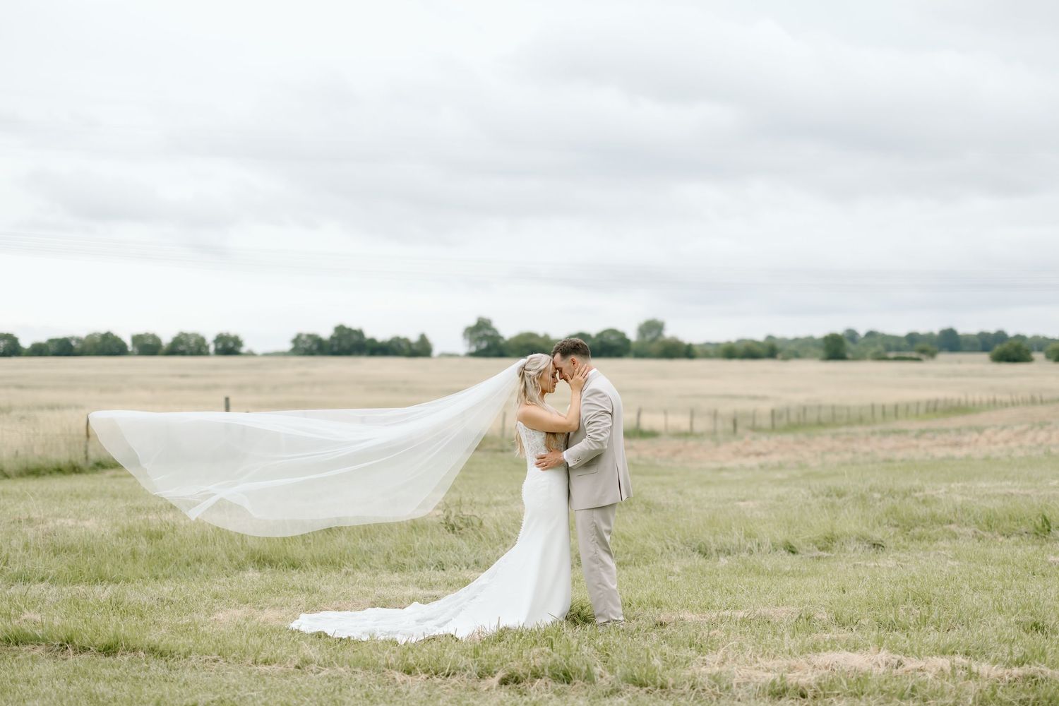 Bride and groom kissing in a green field with veil flowing in the wind on their wedding day.