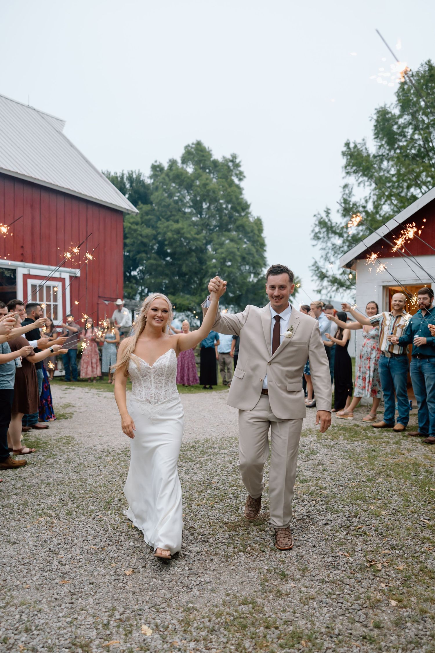 Bride and groom celebrate sparkler exit at rustic barn wedding venue surrounded by cheering guests at dusk.