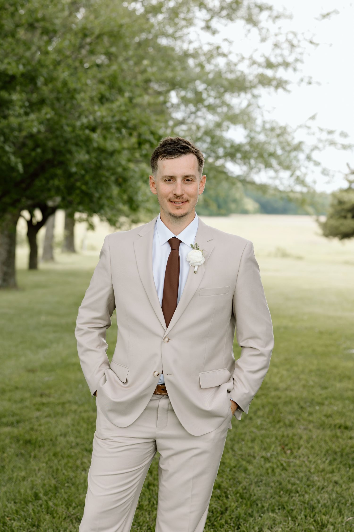 Groom in a beige suit with burgundy tie and white boutonniere stands outdoors on green grass with trees behind him.