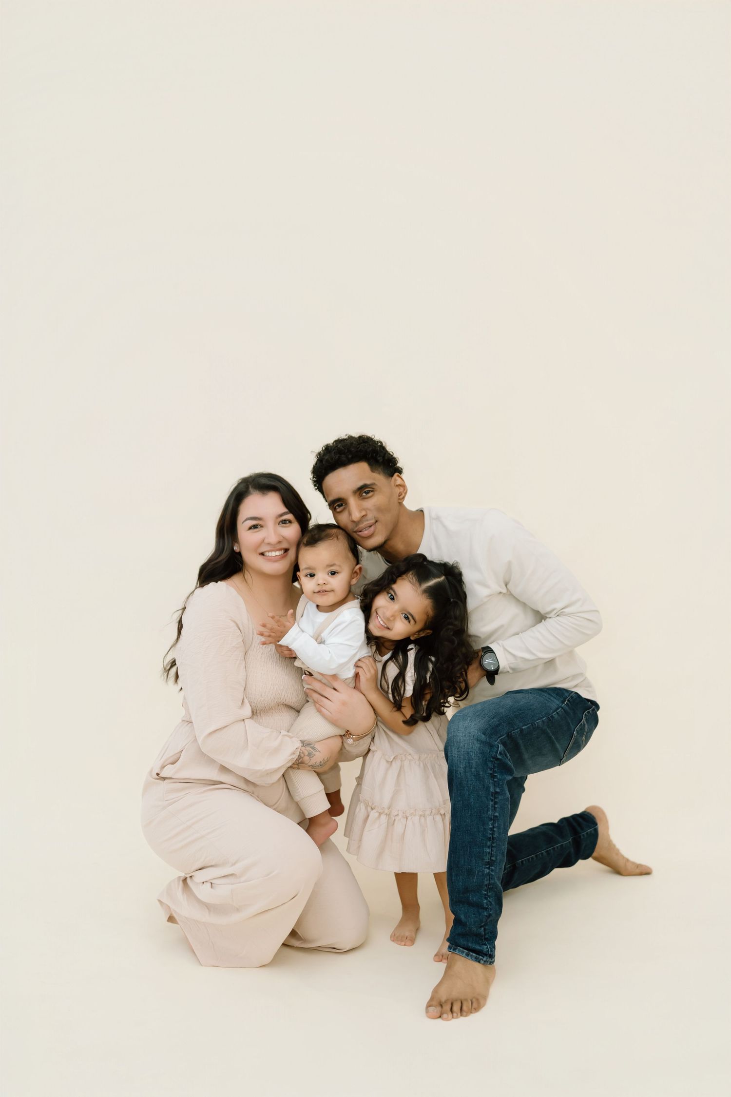 A family wearing neutral colors lies together on a white backdrop in a series of intimate studio portraits.