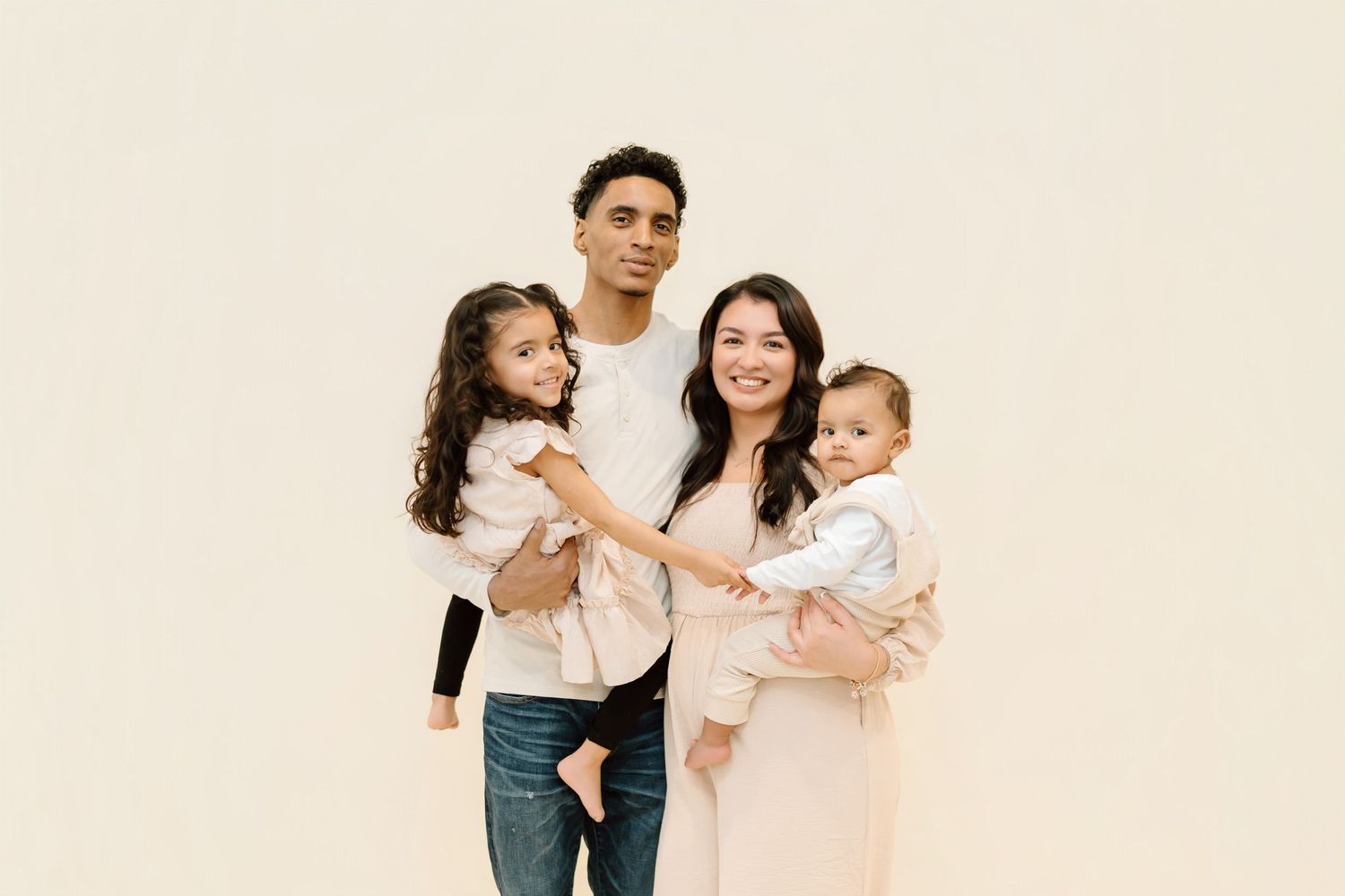 A family in neutral colored clothing poses together against a light beige background in a studio setting.