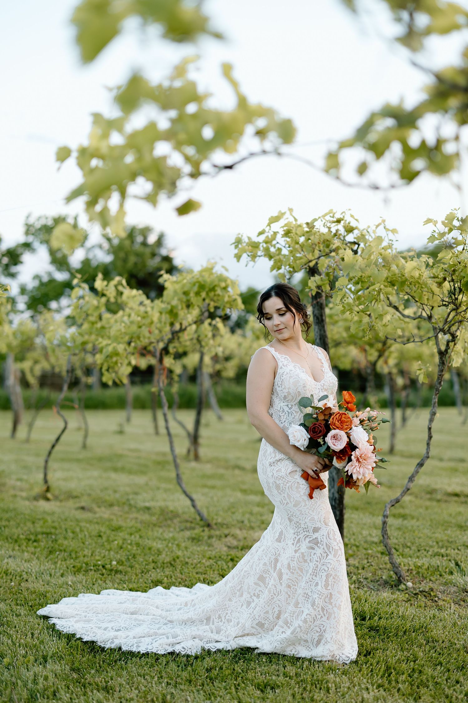 A bride in a lace wedding gown carries a vibrant bouquet while standing in a green orchard setting.