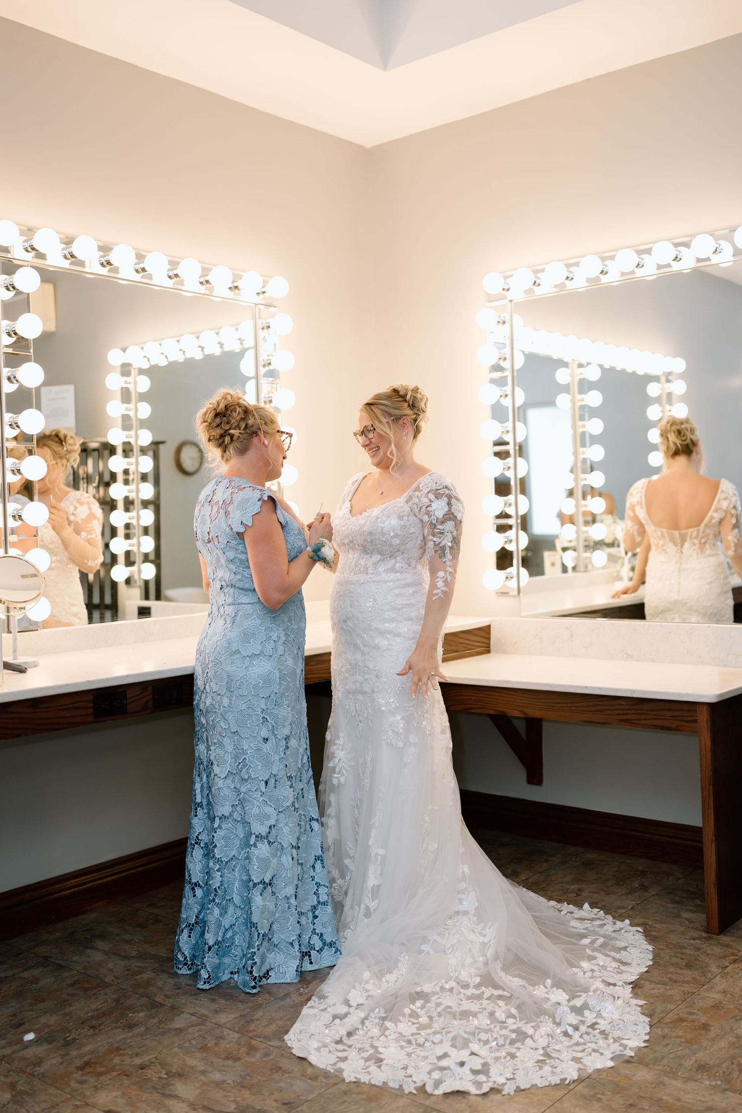 Two people in formal attire stand together in a dressing room with illuminated makeup mirrors.