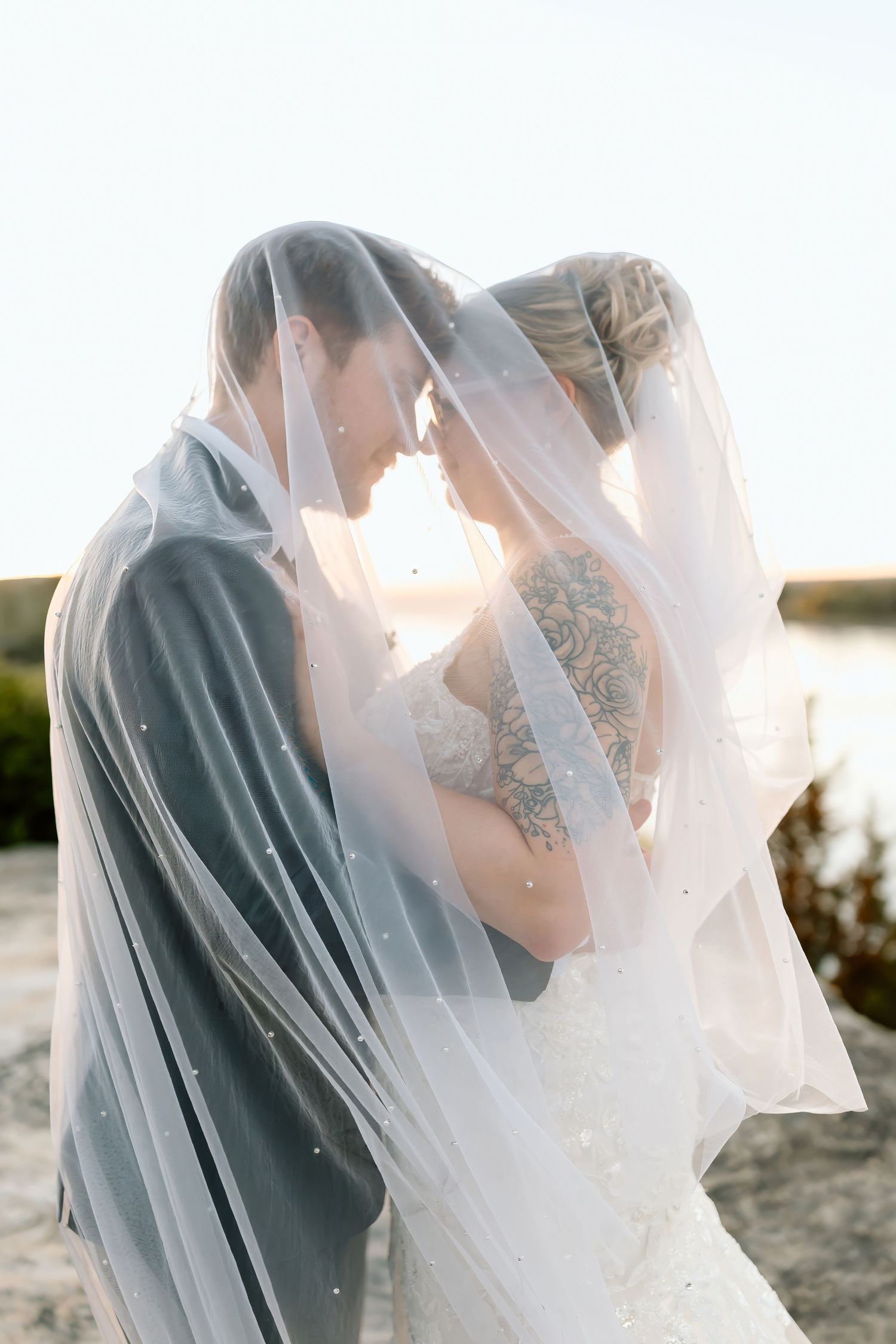 A romantic wedding couple embraces under a flowing white veil during sunset in an outdoor setting.