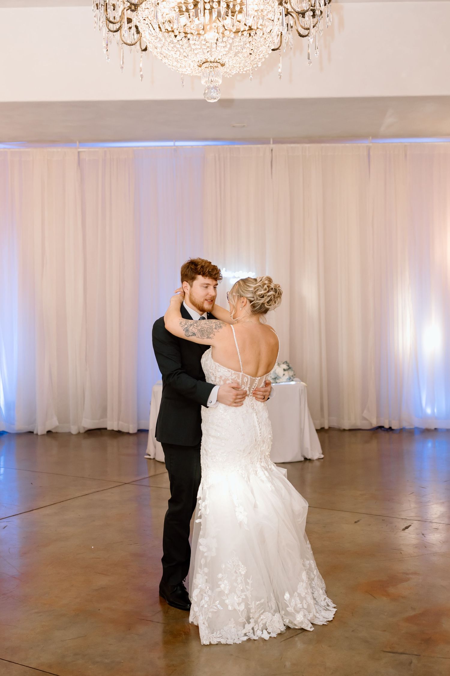 A romantic first dance moment at an elegant wedding reception with white drapes and chandelier backdrop.