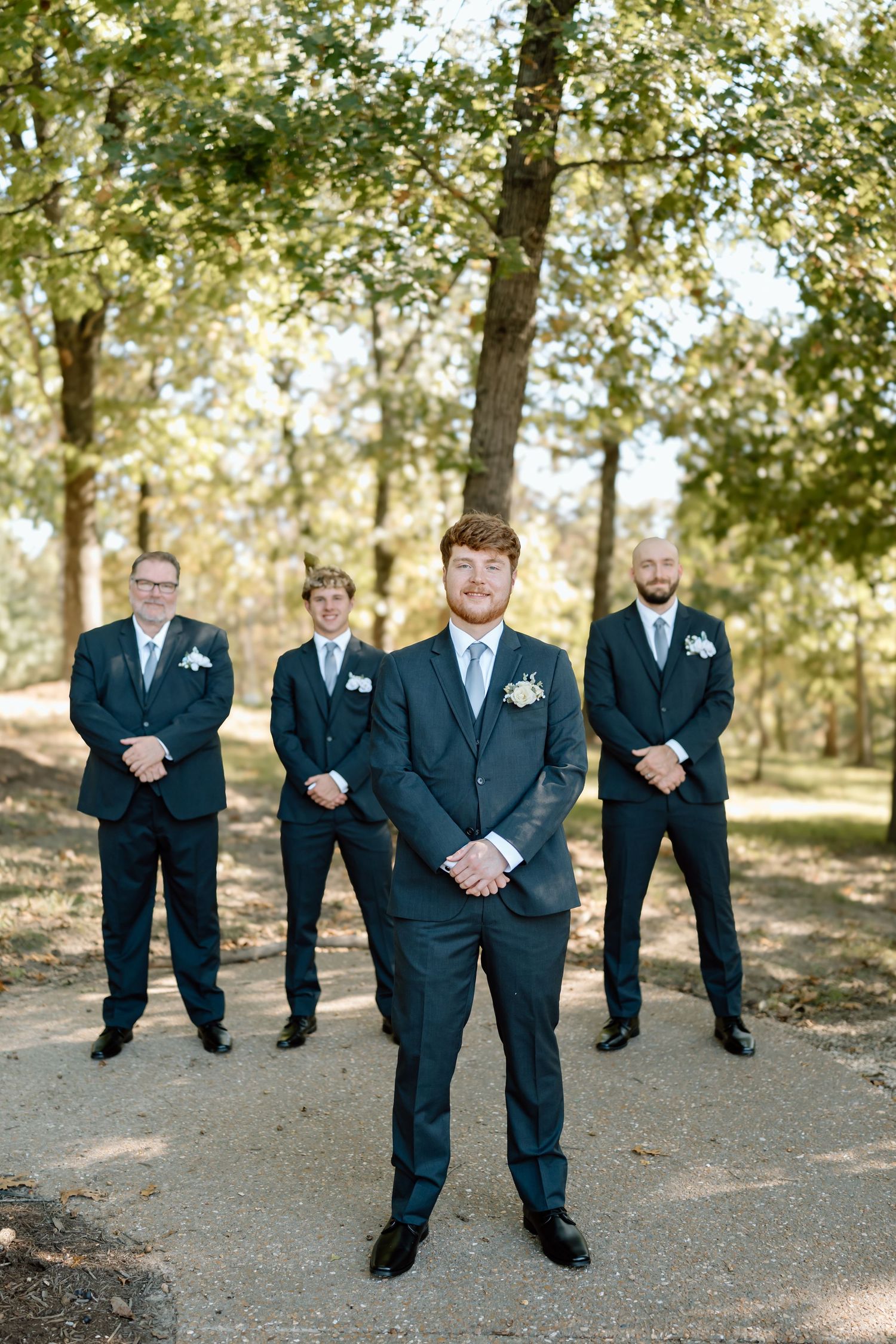 Groomsmen in dark suits stand together outdoors in a wooded area during a wedding photo session.