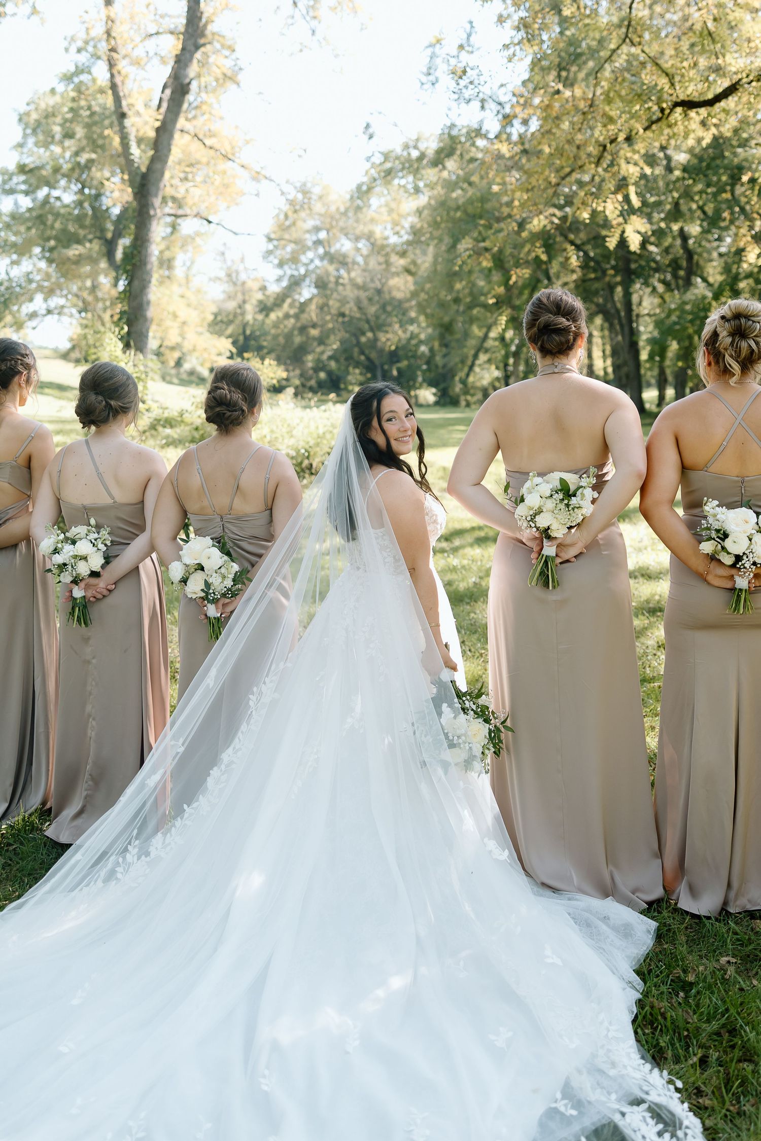Bridal party in champagne colored dresses and white wedding gown stand in line with bouquets at outdoor garden venue.
