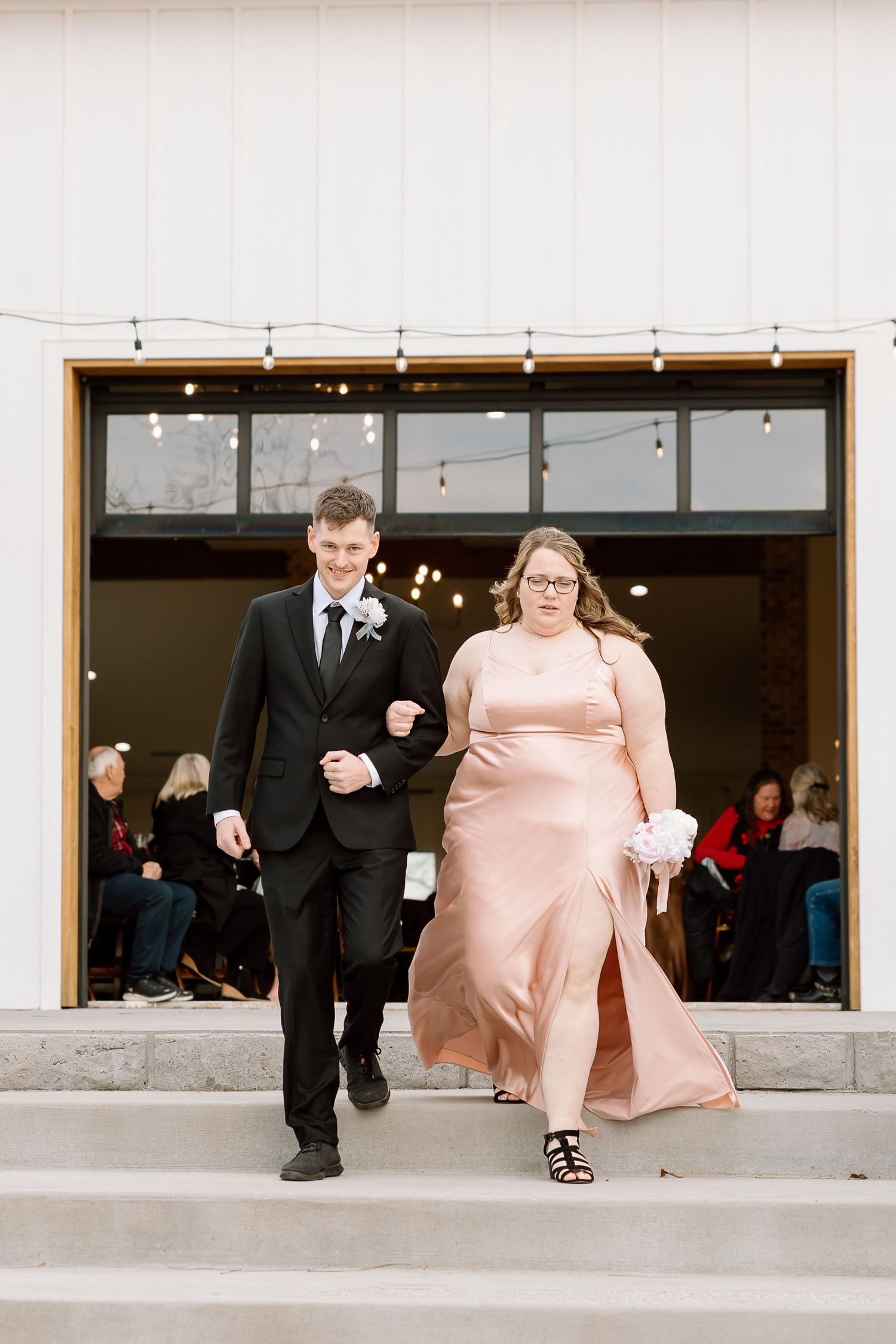 A couple in formal attire walks arm in arm along a concrete walkway with string lights overhead.