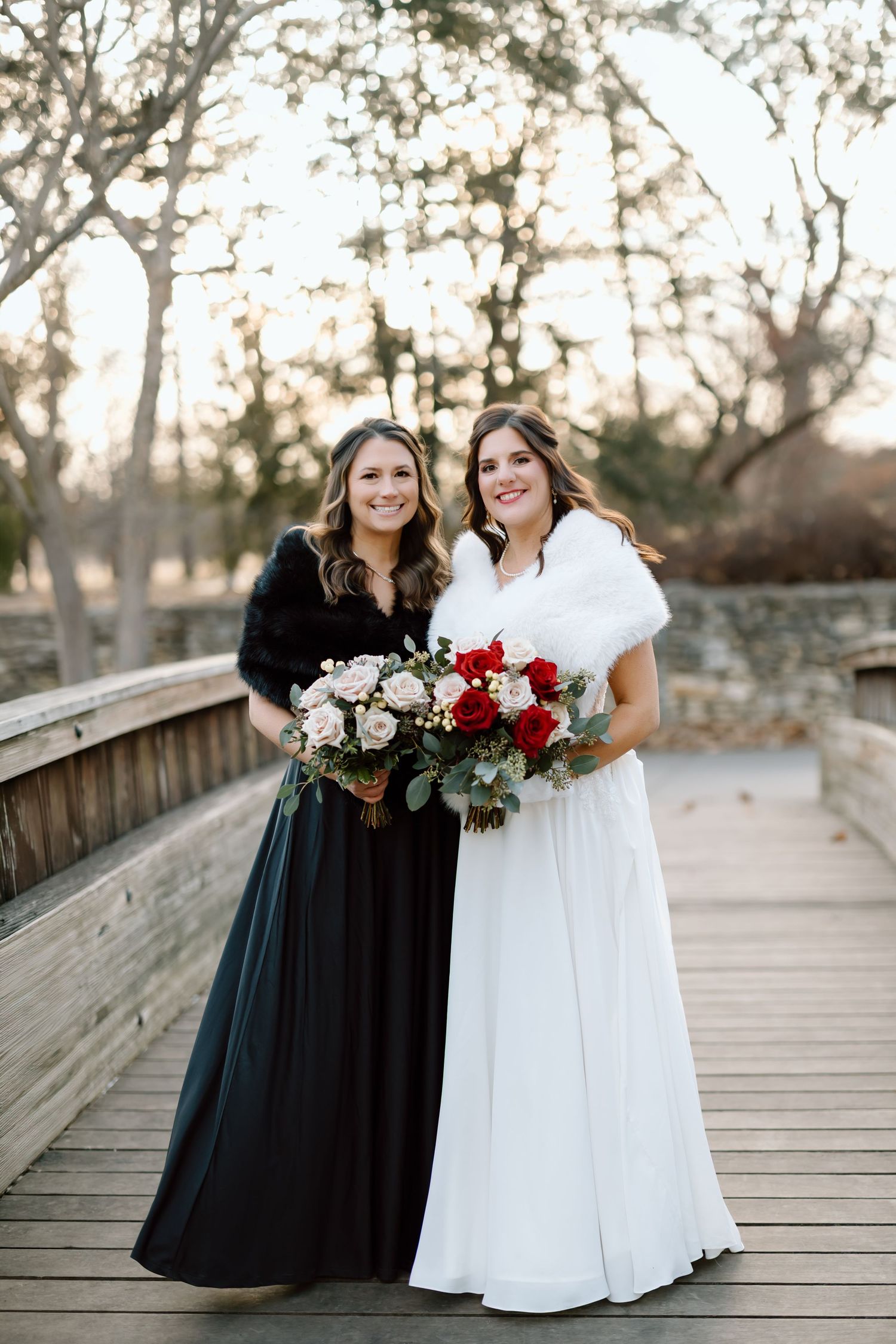 Two people in contrasting black and white dresses pose together on a wooden boardwalk at sunset.