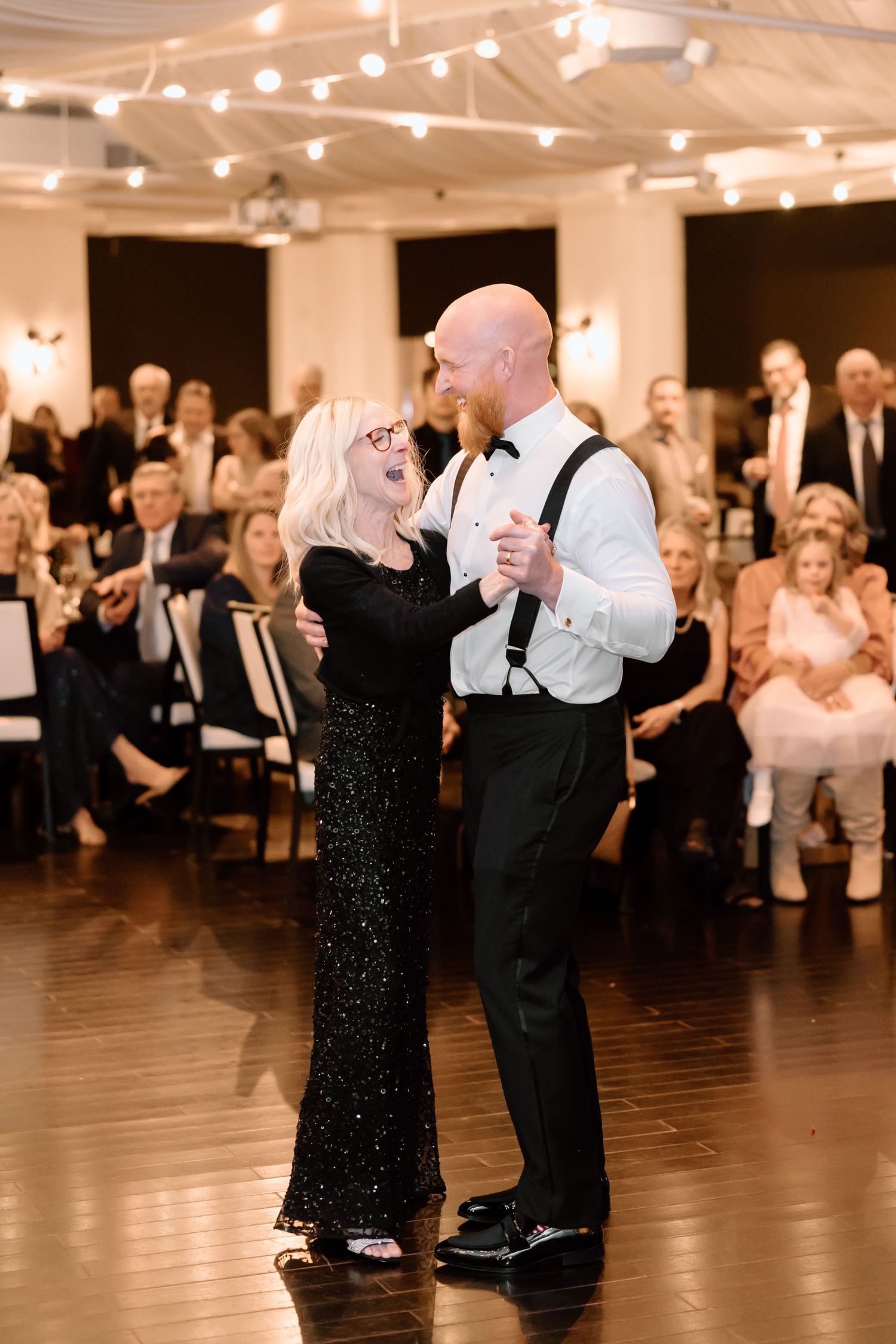Couples dancing together in a spacious ballroom with warm lighting and onlookers in the background.