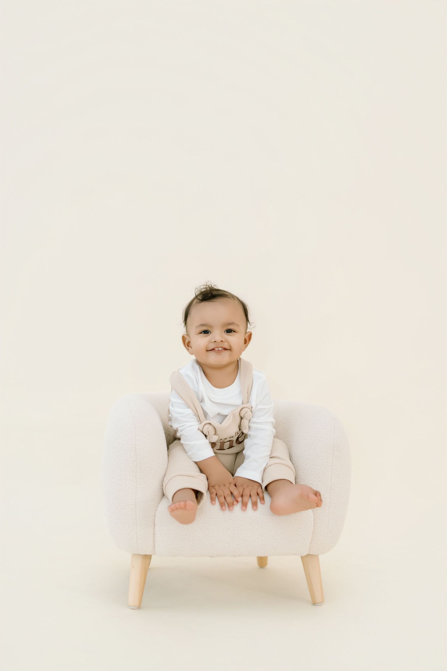 Baby in white outfit sits playfully on a modern white chair against neutral background.