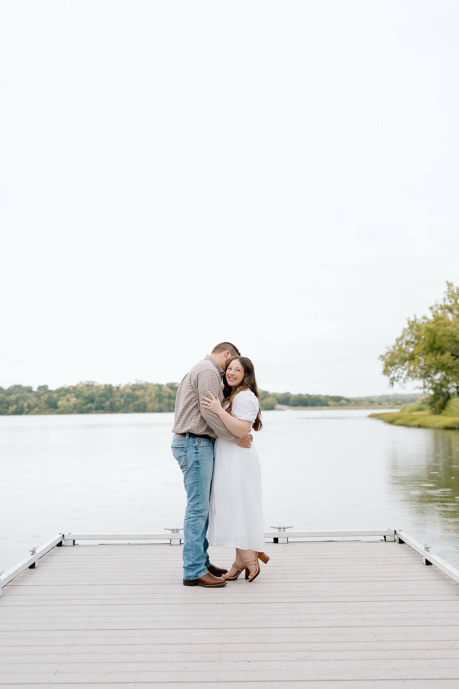A loving couple holds each other close while standing on a dock by the water.