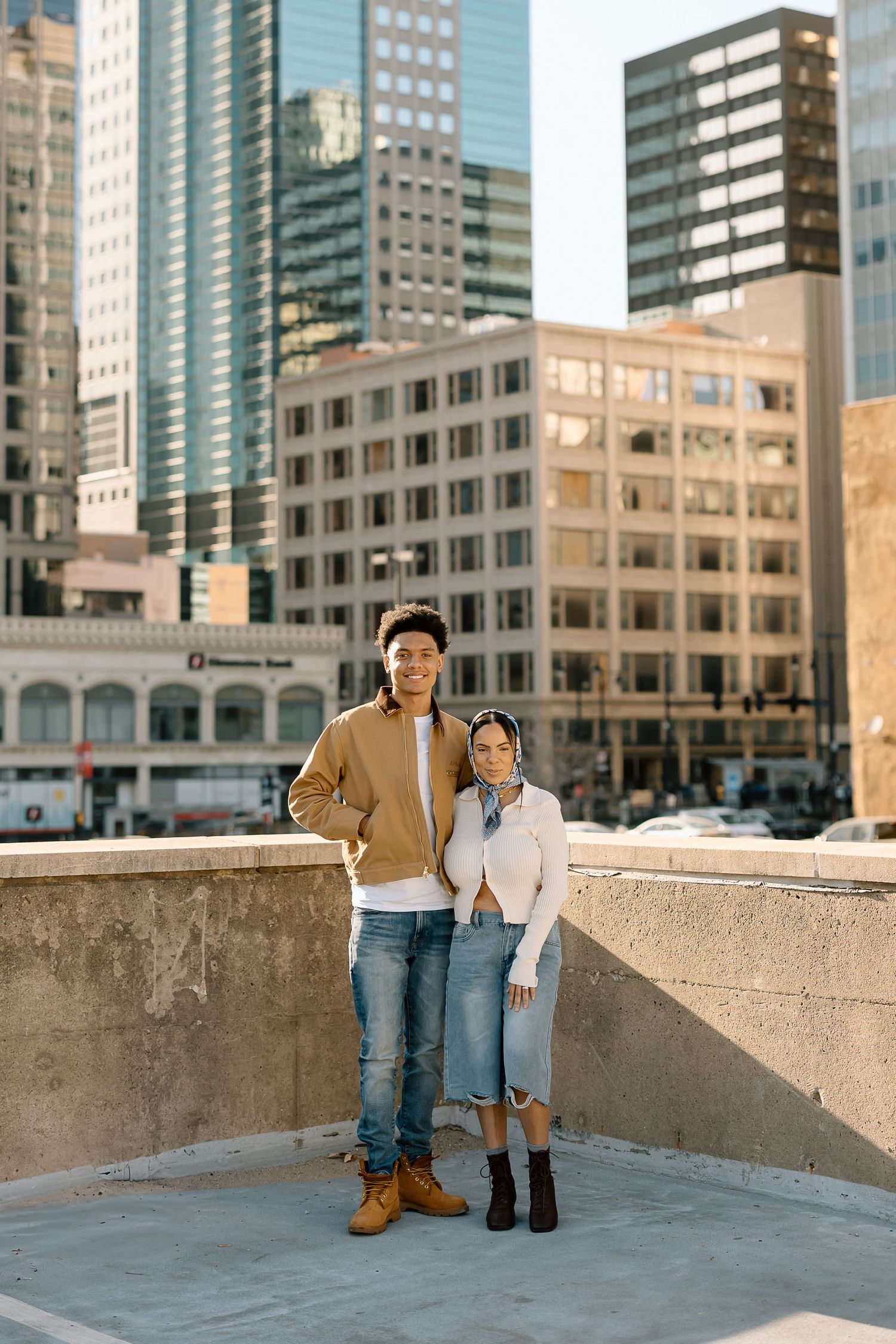 A couple poses on a rooftop with urban skyscrapers in the background on a sunny day.