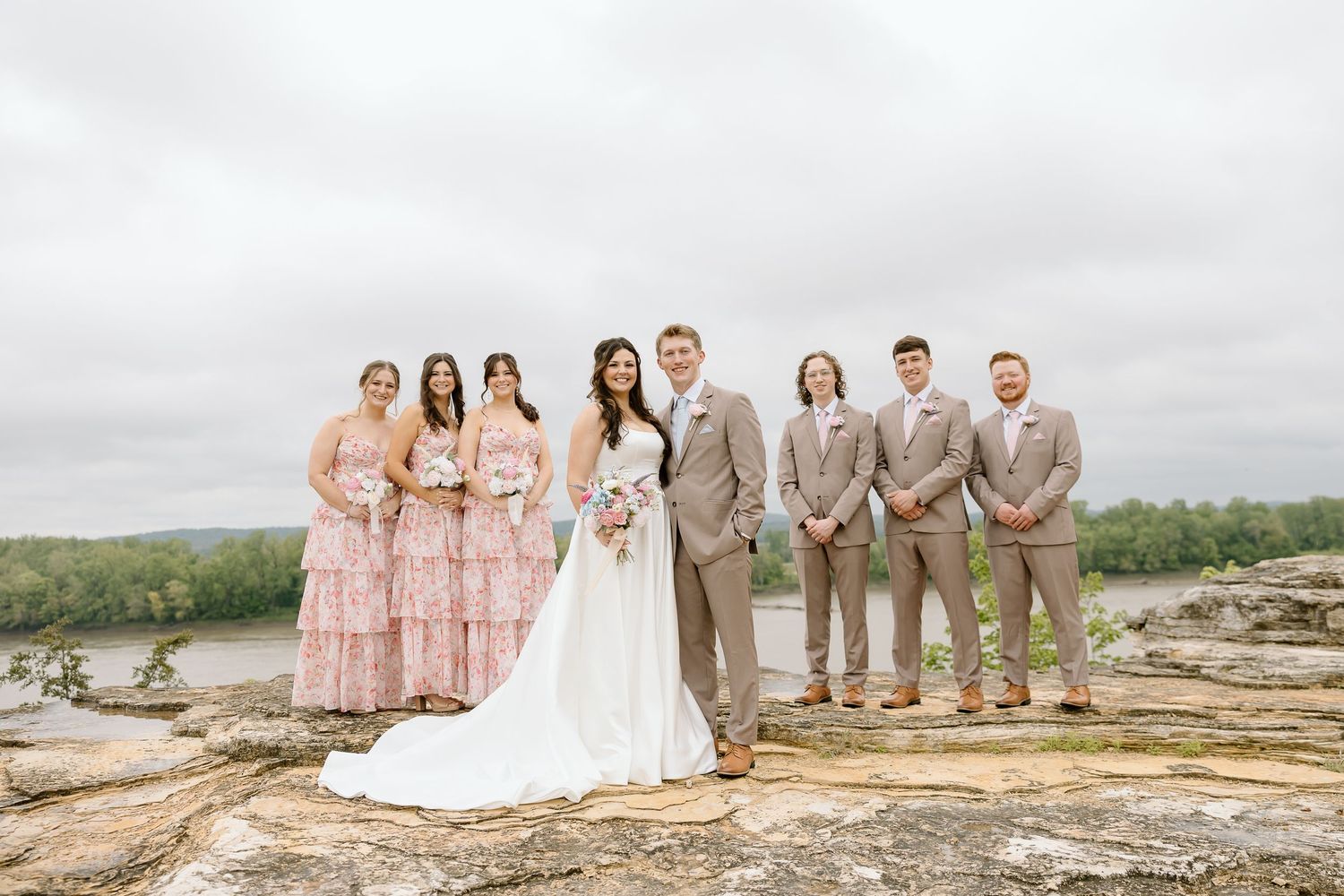 Wedding party in blush pink dresses and tan suits gathered on rocky overlook with scenic background.