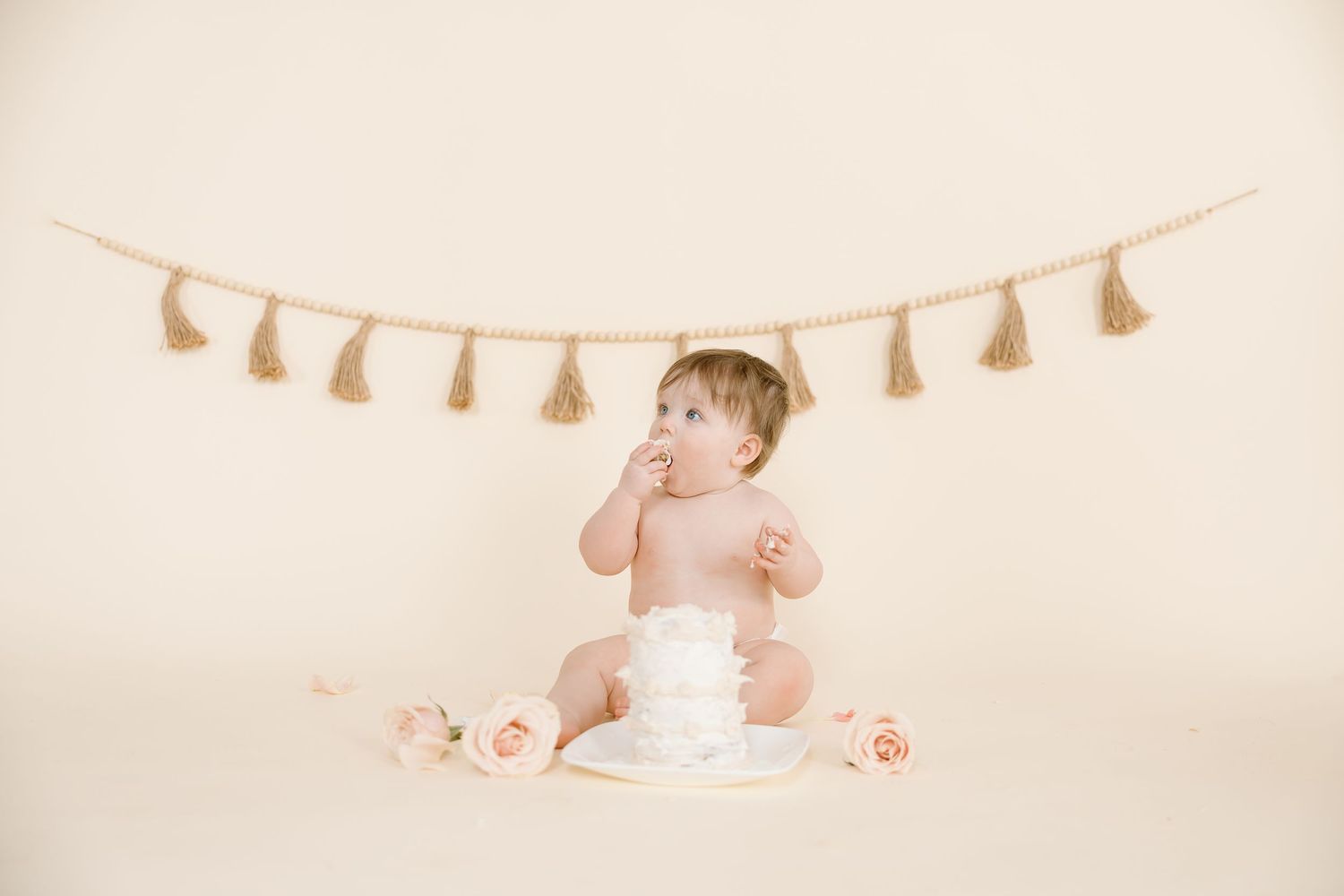 A baby touching face near white cake with pink roses and tassel garland above.