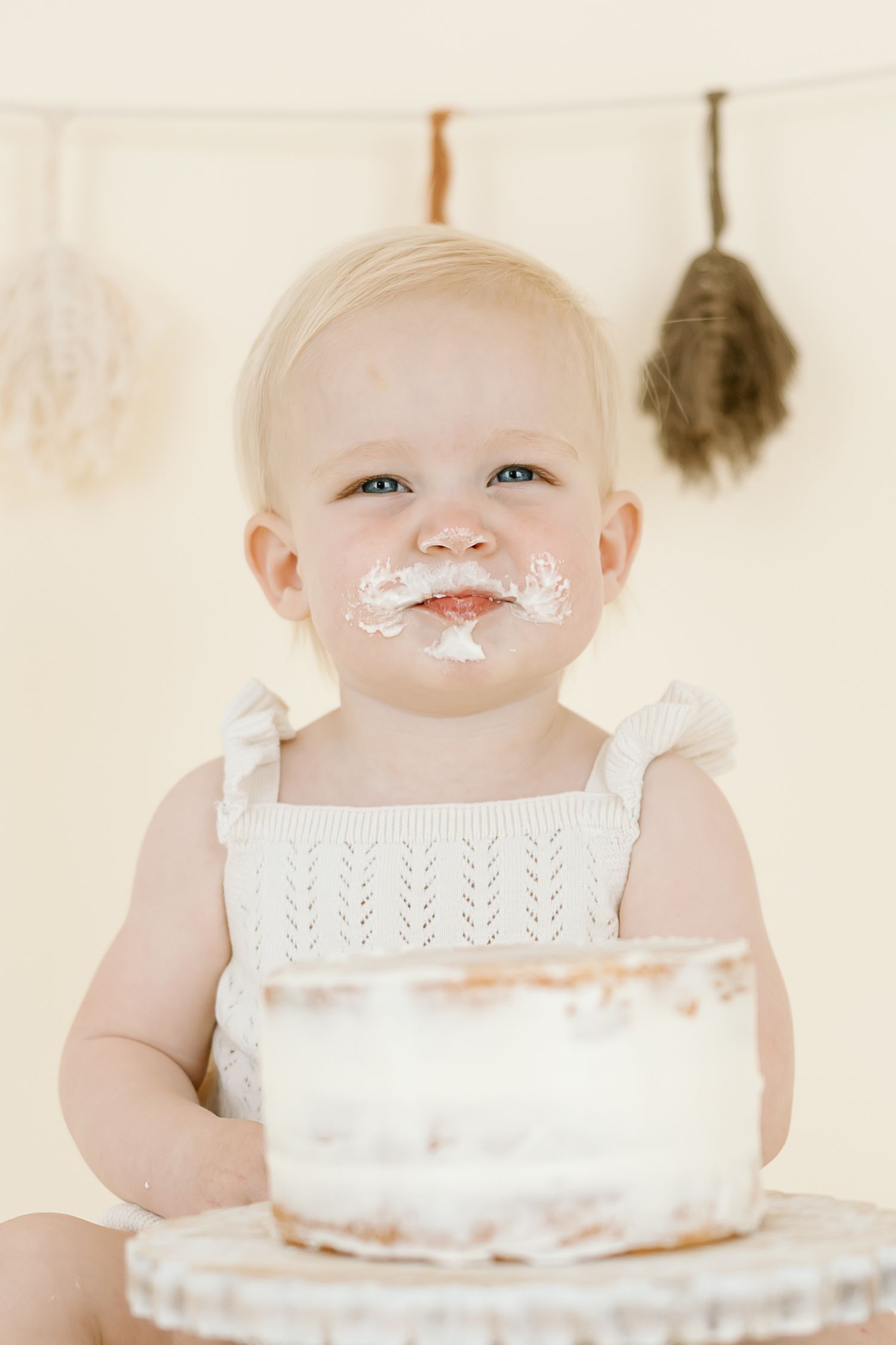 A baby wearing a white bib smiles while playing with cake frosting and making a delightful mess during a cake smash photoshoot.