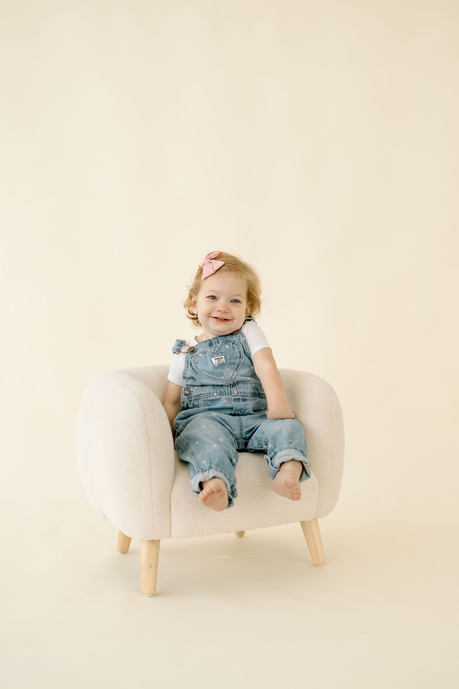 Small child wearing denim overalls sits in a modern white chair against neutral background.