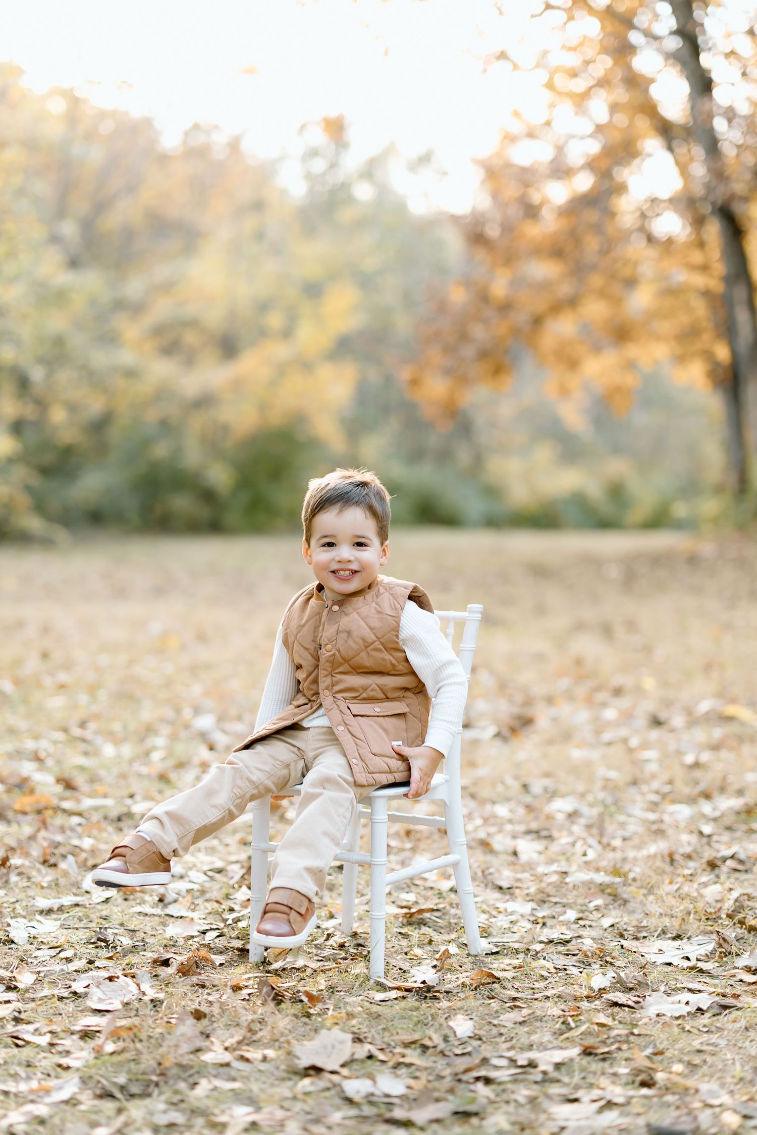 A child sits in a white Adirondack chair surrounded by fallen autumn leaves in a park setting.