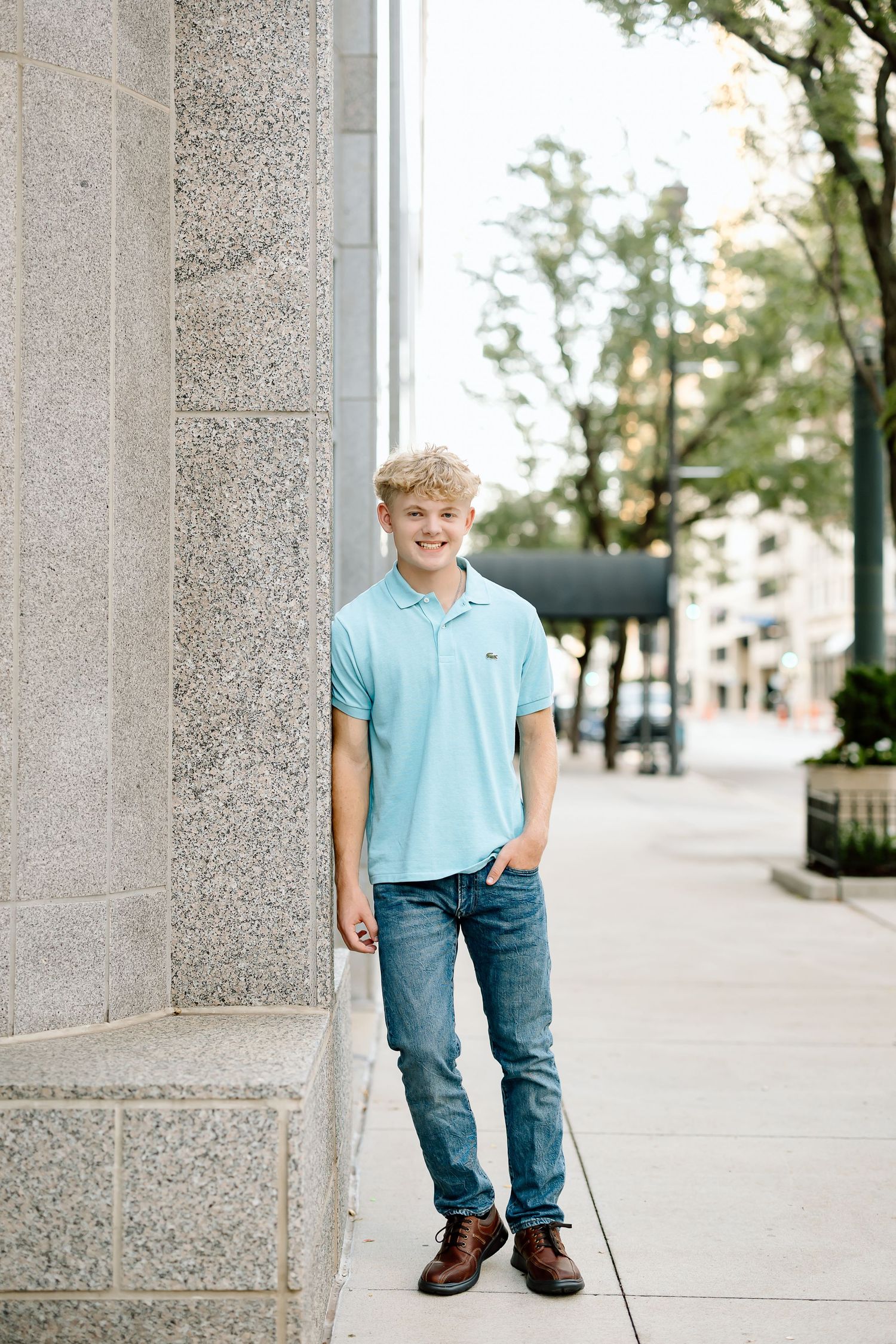 Young person in light blue polo shirt and jeans standing casually against stone column on city sidewalk.