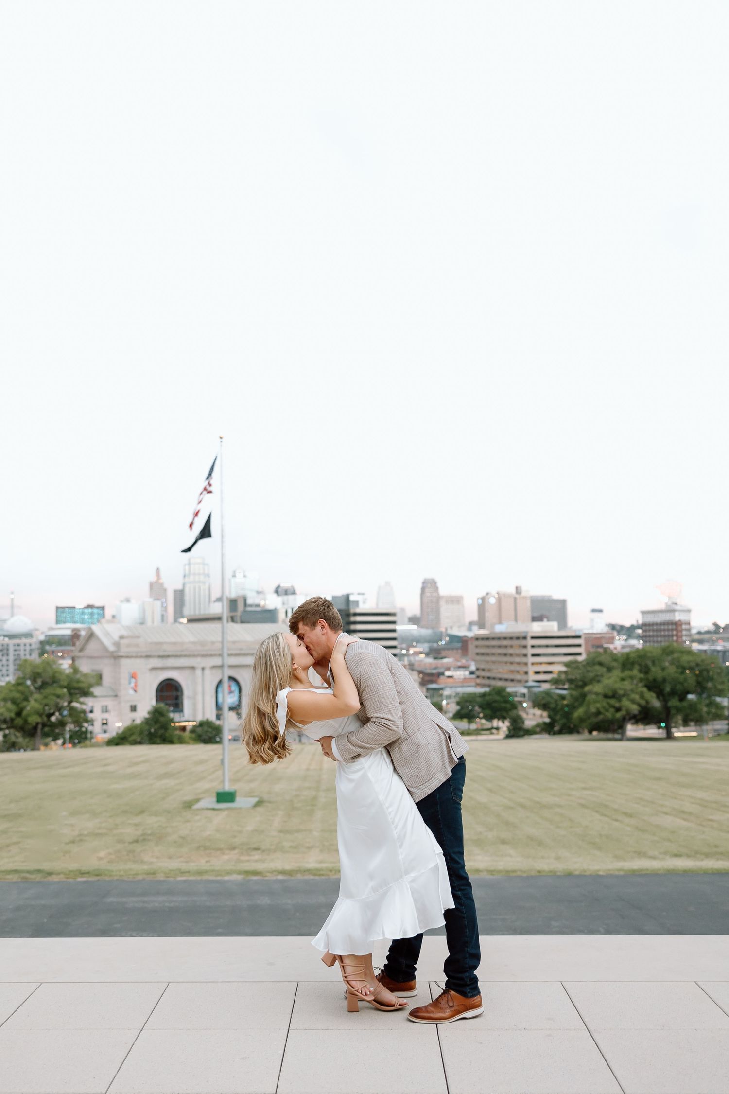 A romantic couple in wedding attire share an intimate dip pose on a city rooftop with an urban skyline in the background.