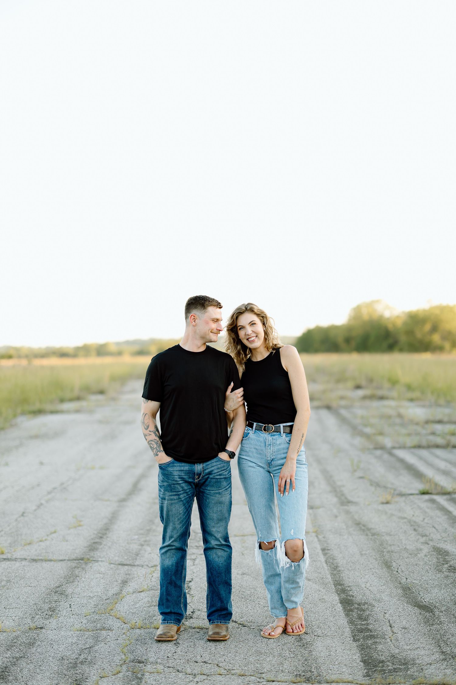 A couple wearing black tops and blue jeans stands together on a dirt road at sunset.