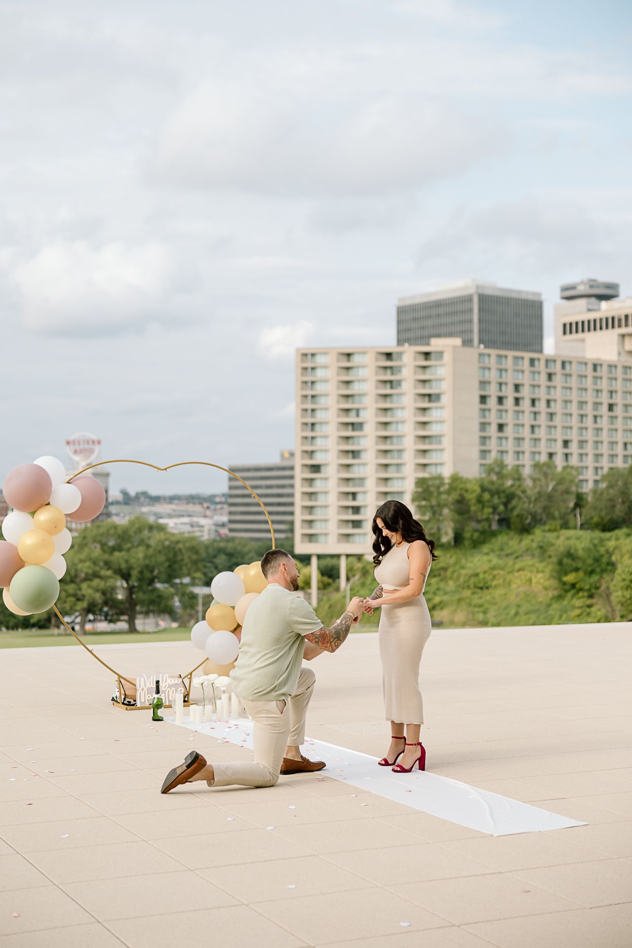 A romantic proposal setup on a rooftop with pastel balloons and city skyline in the background.