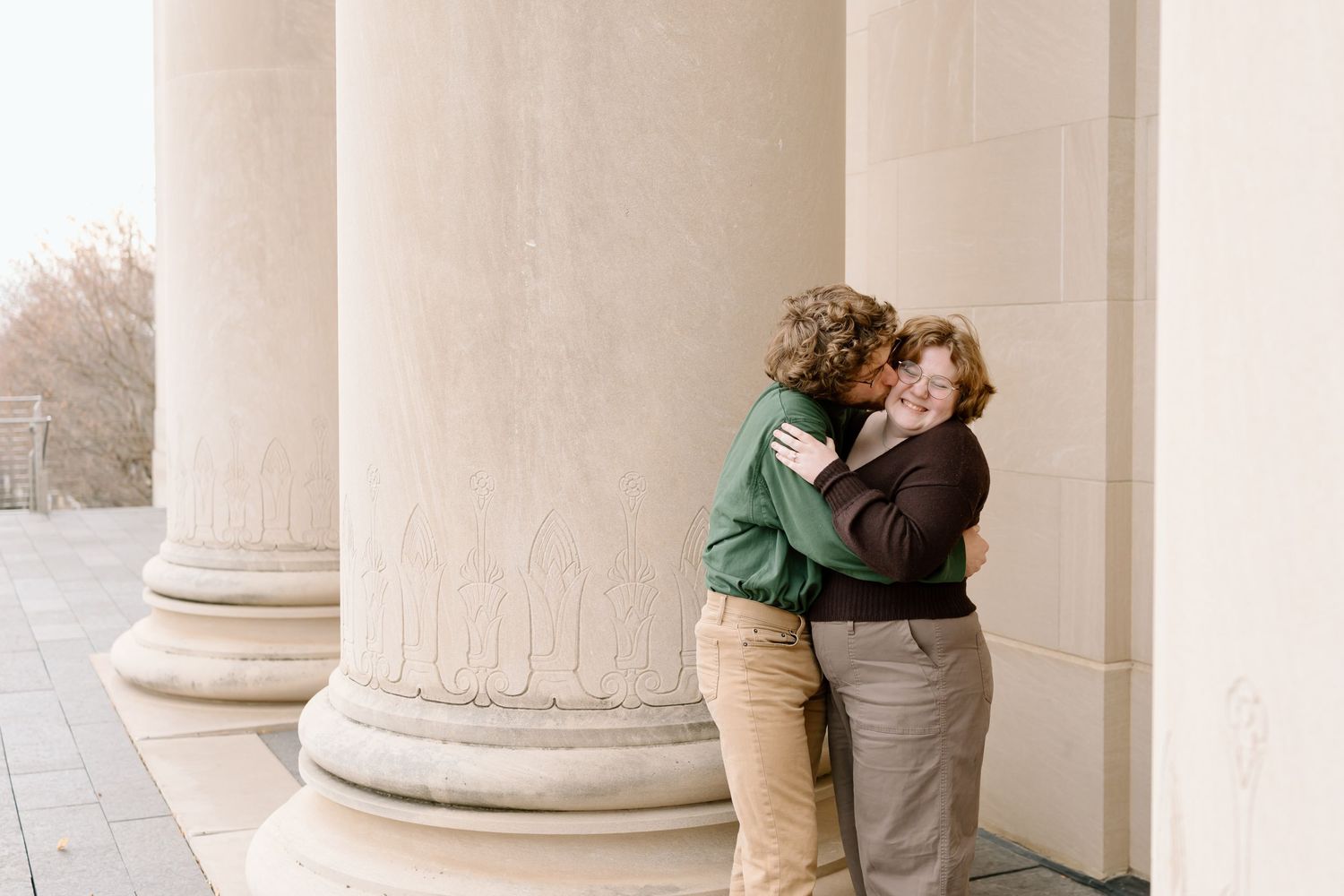 Two people embrace in a tender moment between classical stone columns in warm, muted tones.