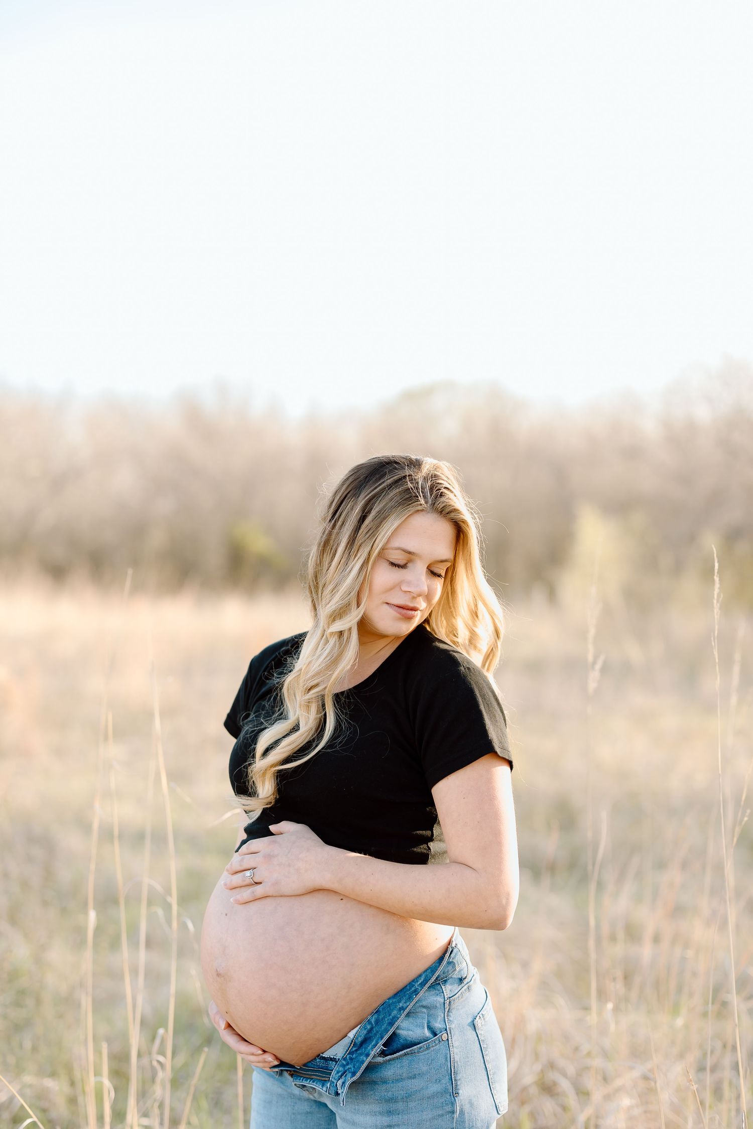 Expectant mother in black top and jeans embraces her belly while standing in a sunlit field of tall grass.