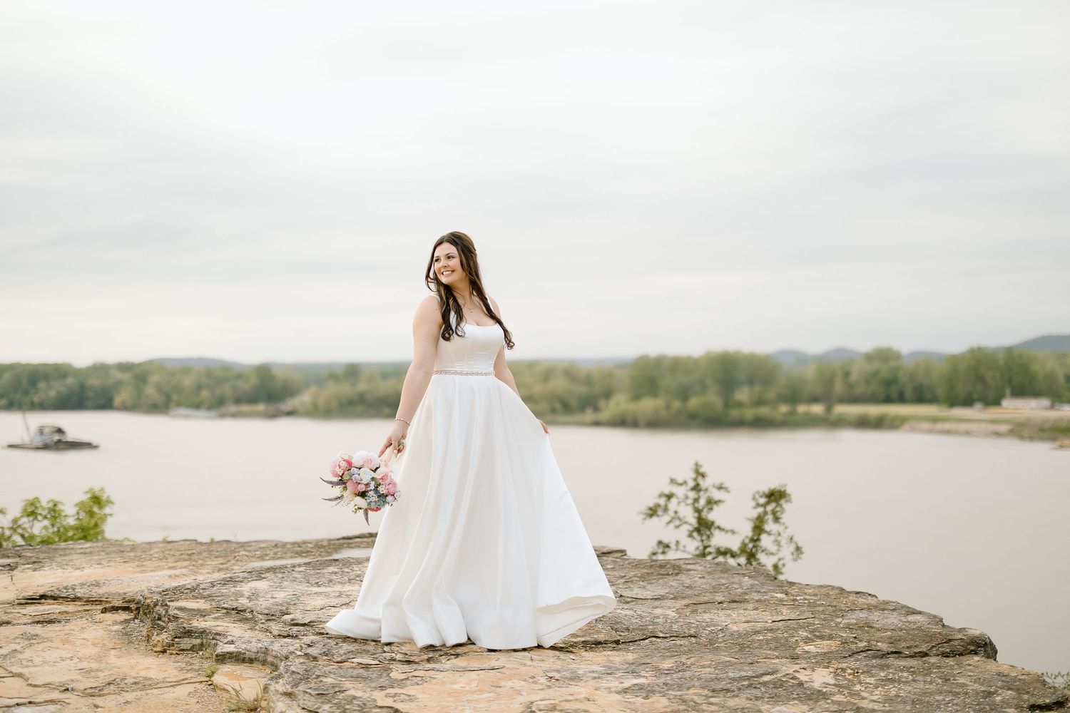 A person in a flowing white wedding dress stands on a rocky overlook by a serene river on an overcast day.