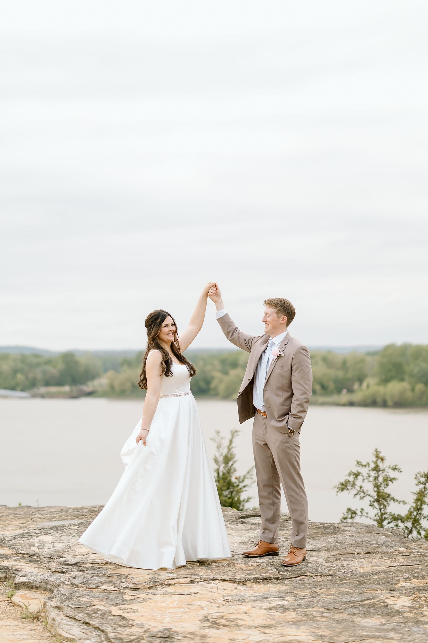 A couple in wedding attire dances together on a rocky overlook by a serene lake on their special day.
