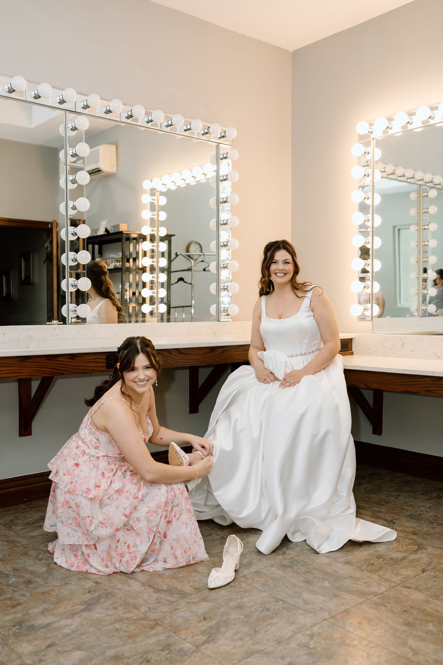 Two people in white and pink dresses sit together in front of a vanity mirror with bright makeup lights.