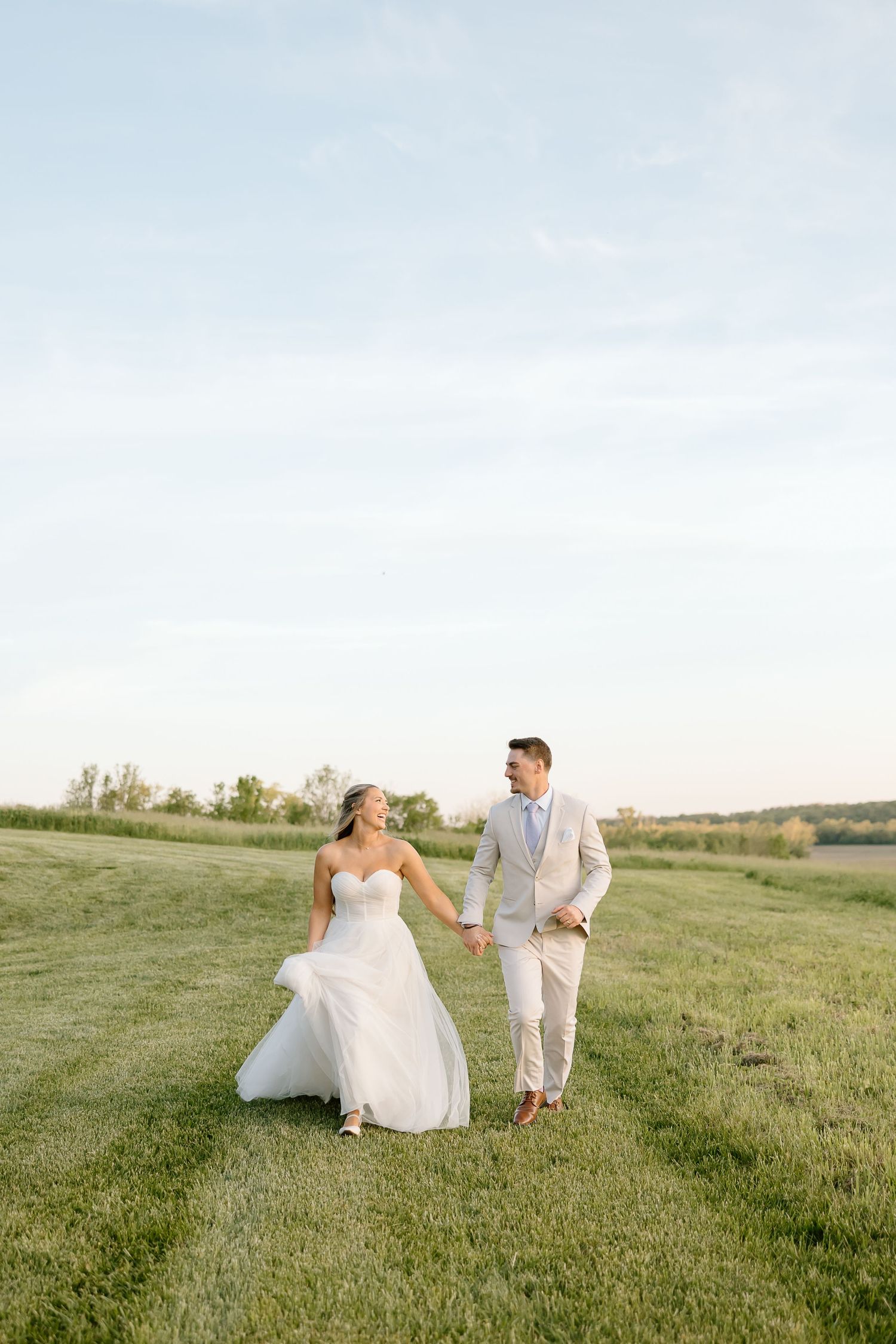 A bride and groom walk together across a scenic grassy hilltop during their outdoor wedding photoshoot at sunset.
