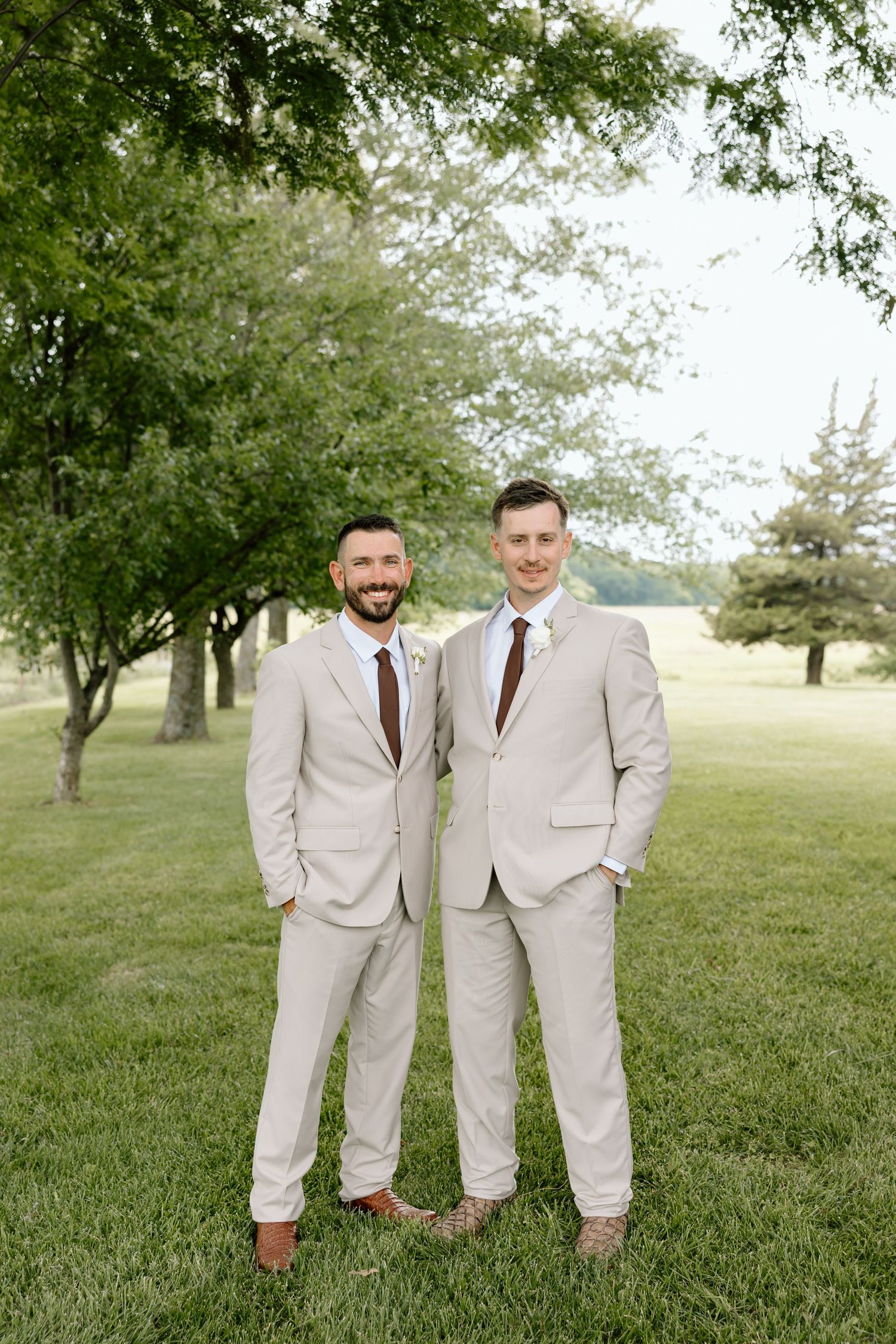 Two grooms in matching tan suits and brown ties smile together outdoors on a green lawn at their wedding.