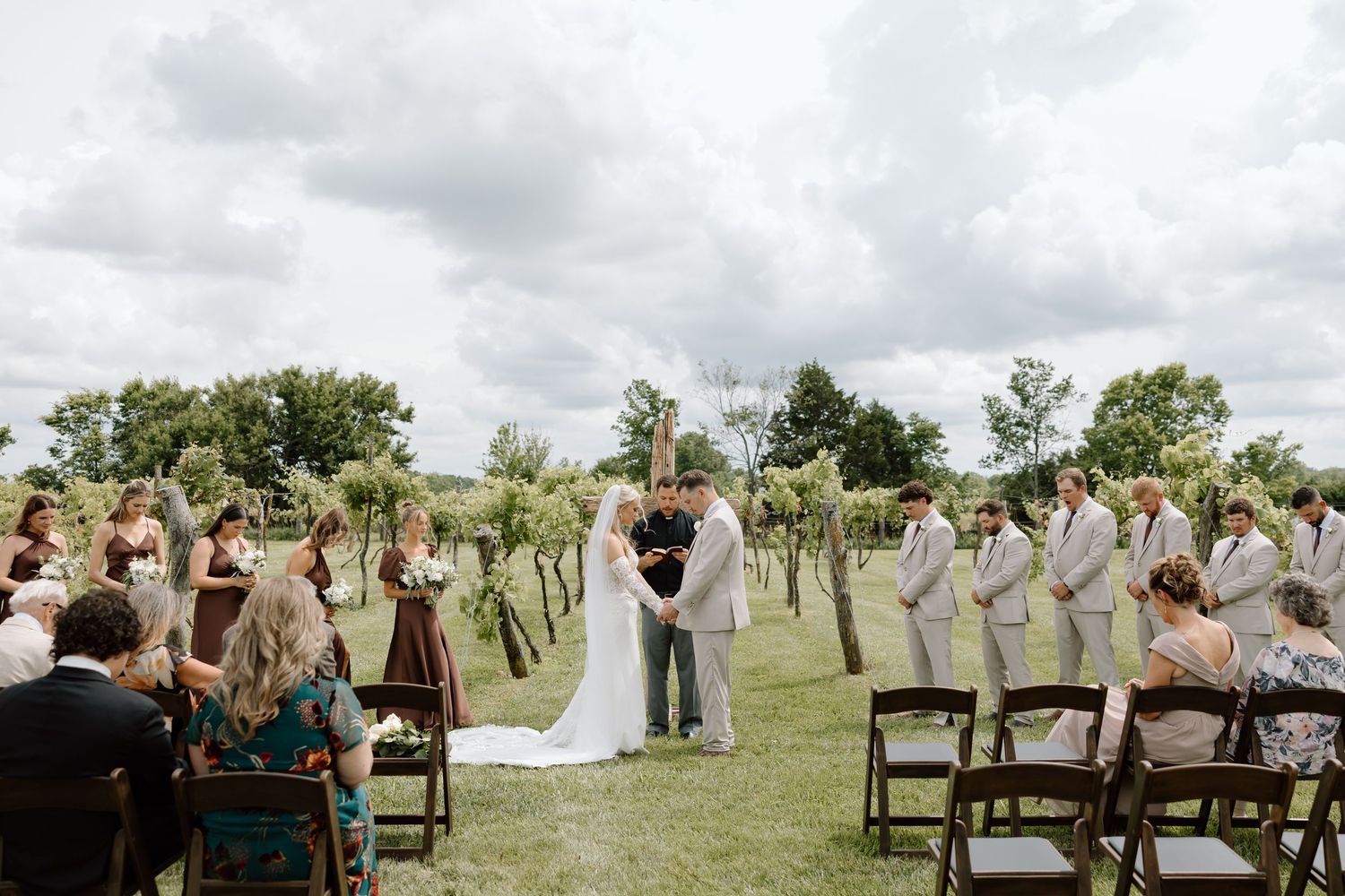 Outdoor vineyard wedding ceremony with bride and groom at altar, bridesmaids in burgundy, groomsmen in gray, under cloudy skies.