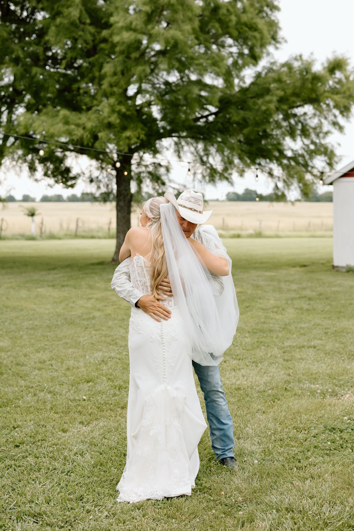 A bride in a lace gown and veil shares a romantic kiss with a groom in a cowboy hat and jeans outdoors.