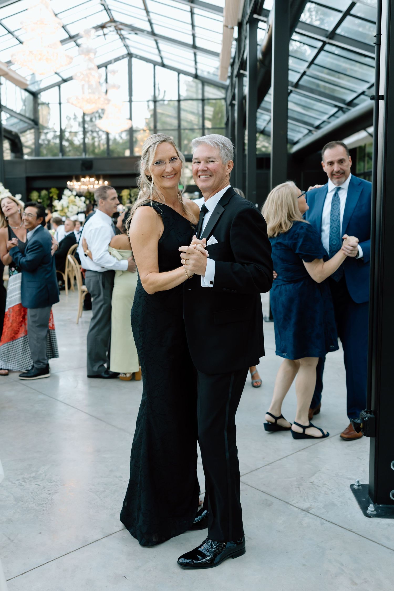 Elegant couple dancing in black formal attire at a wedding reception in a glass greenhouse venue.