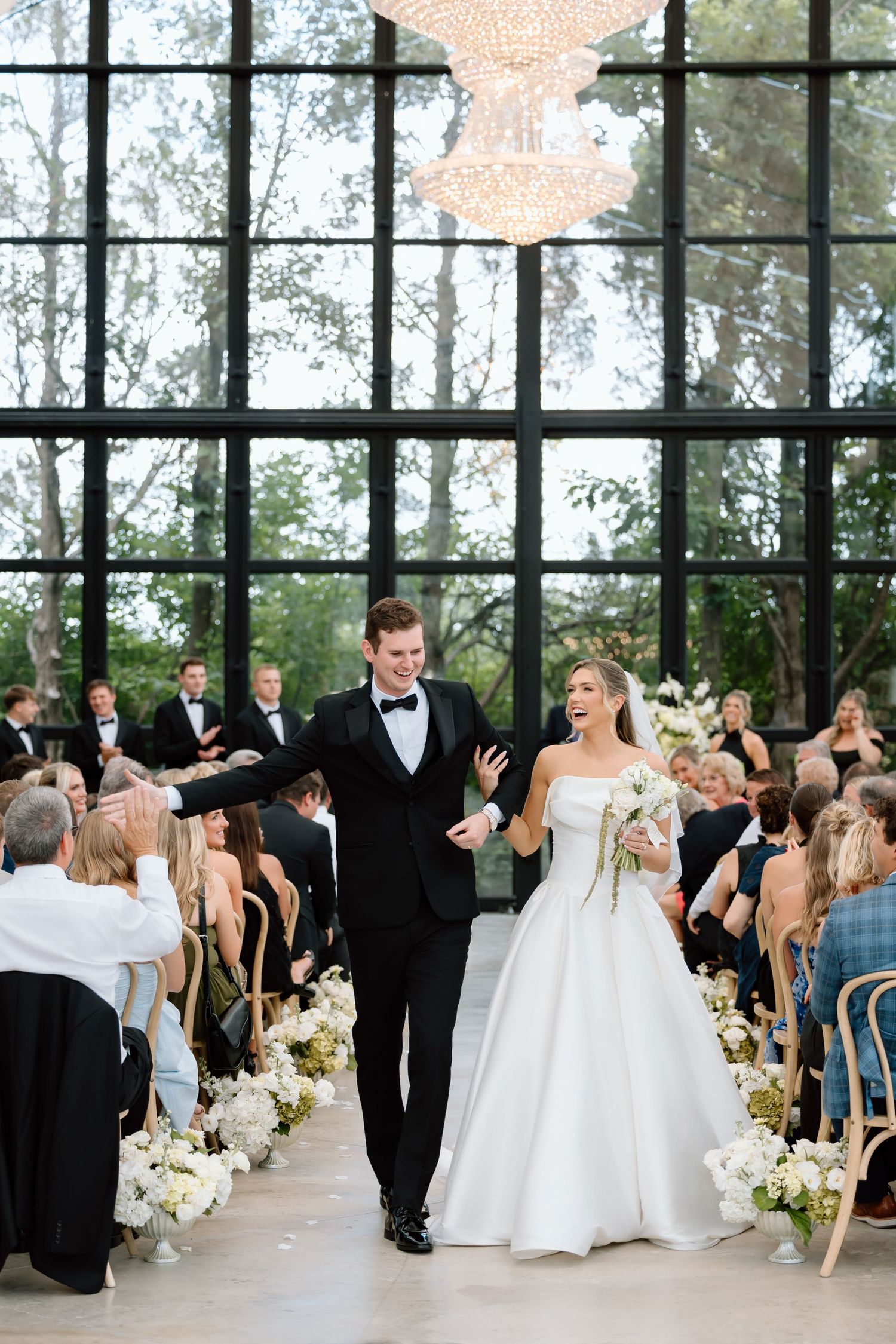 Joyful newlyweds walk down the aisle in an elegant glass-walled venue, bride in white gown holding bouquet.