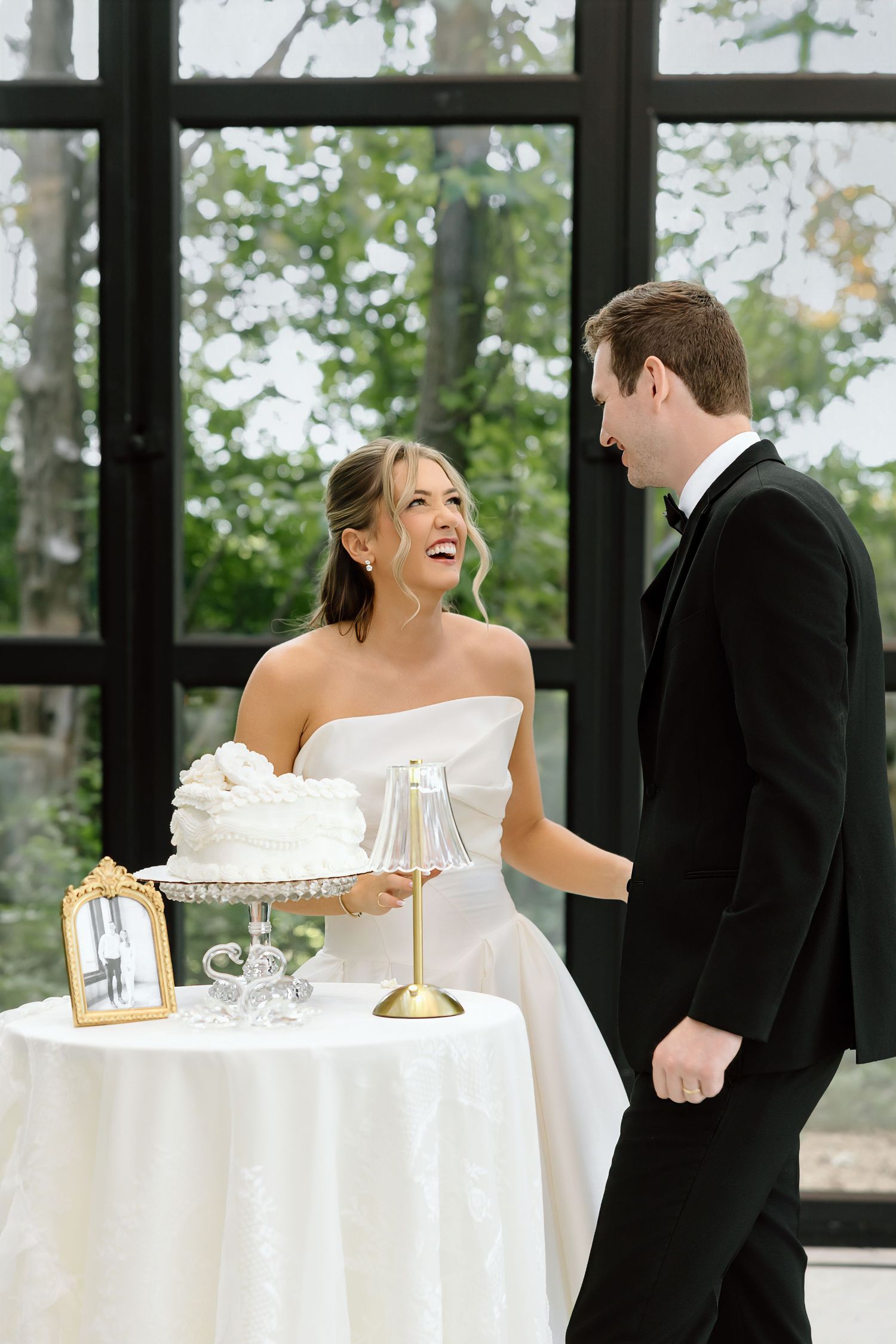 Bride and groom laughing together beside a white wedding cake on a draped table in an elegant glass-windowed venue.