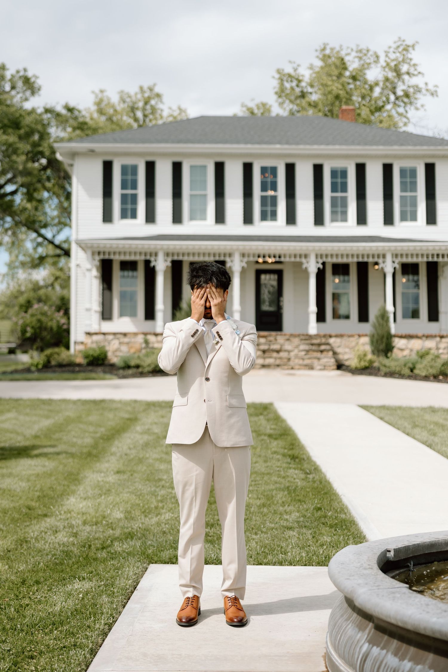 Groom in cream suit covering face with hands, standing on pathway before elegant white colonial mansion.