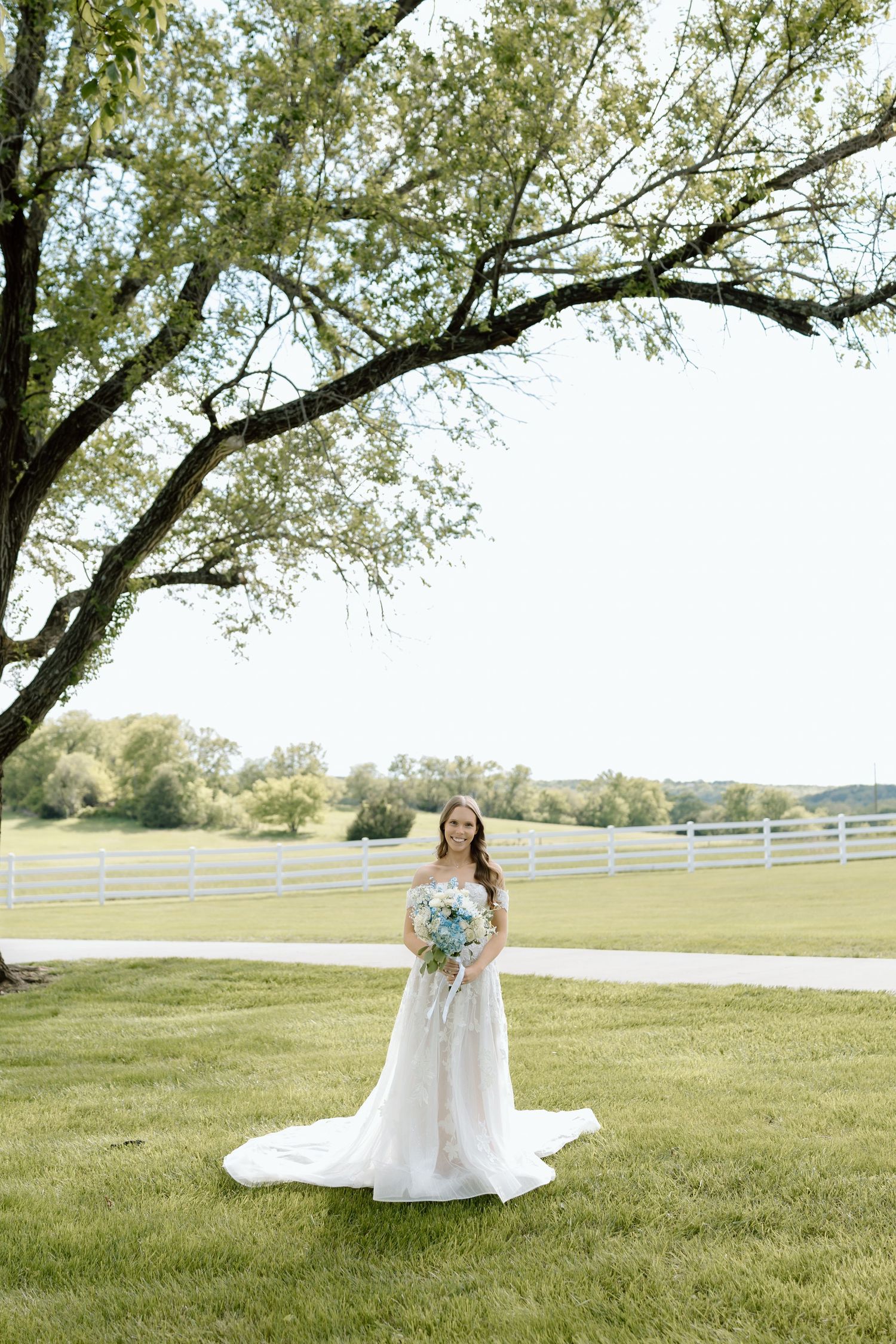 Bride in white off-shoulder gown holding blue bouquet stands under large tree on green lawn with white fence.