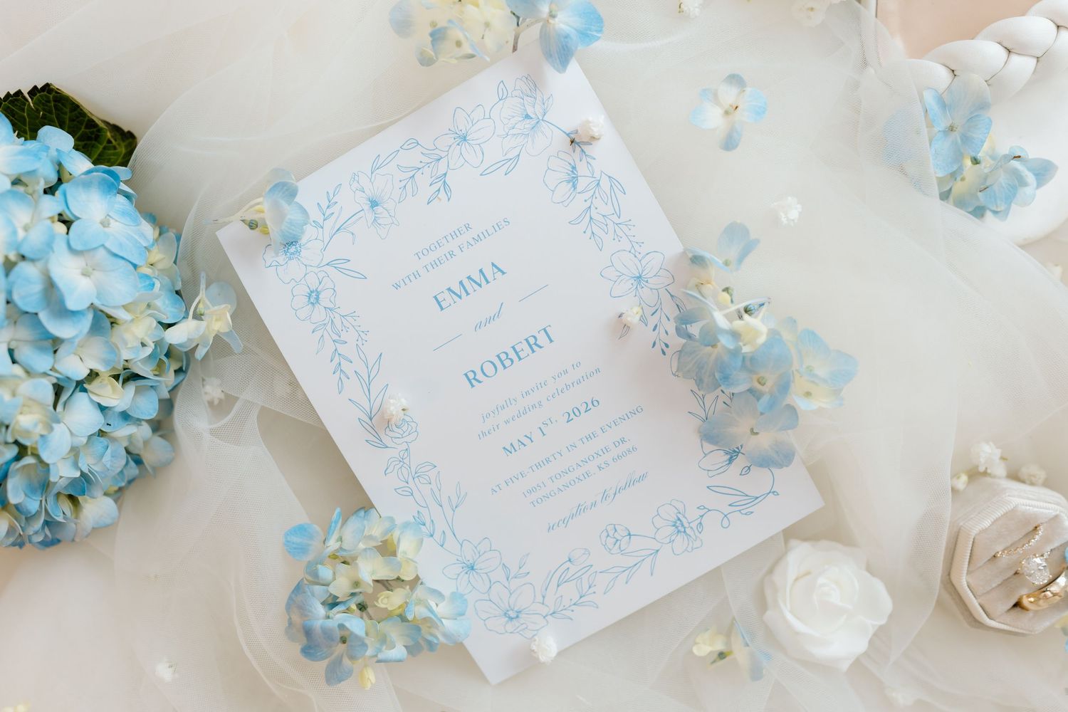 Elegant wedding invitation for Emma and Robert surrounded by blue hydrangeas and white roses on a soft white background.