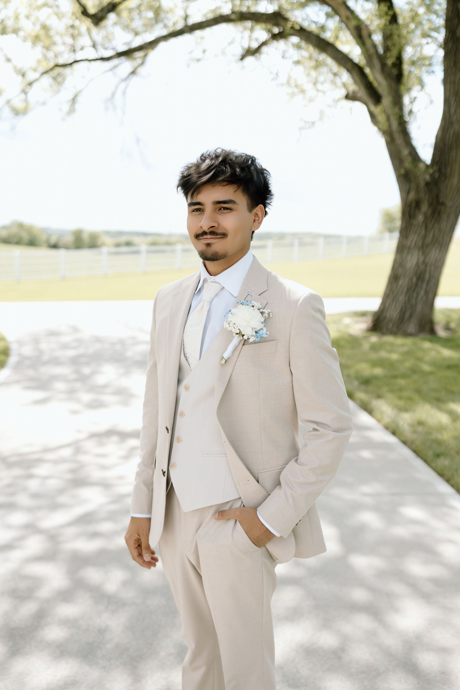 Groom in beige suit with boutonniere poses outdoors near tree on wedding day.